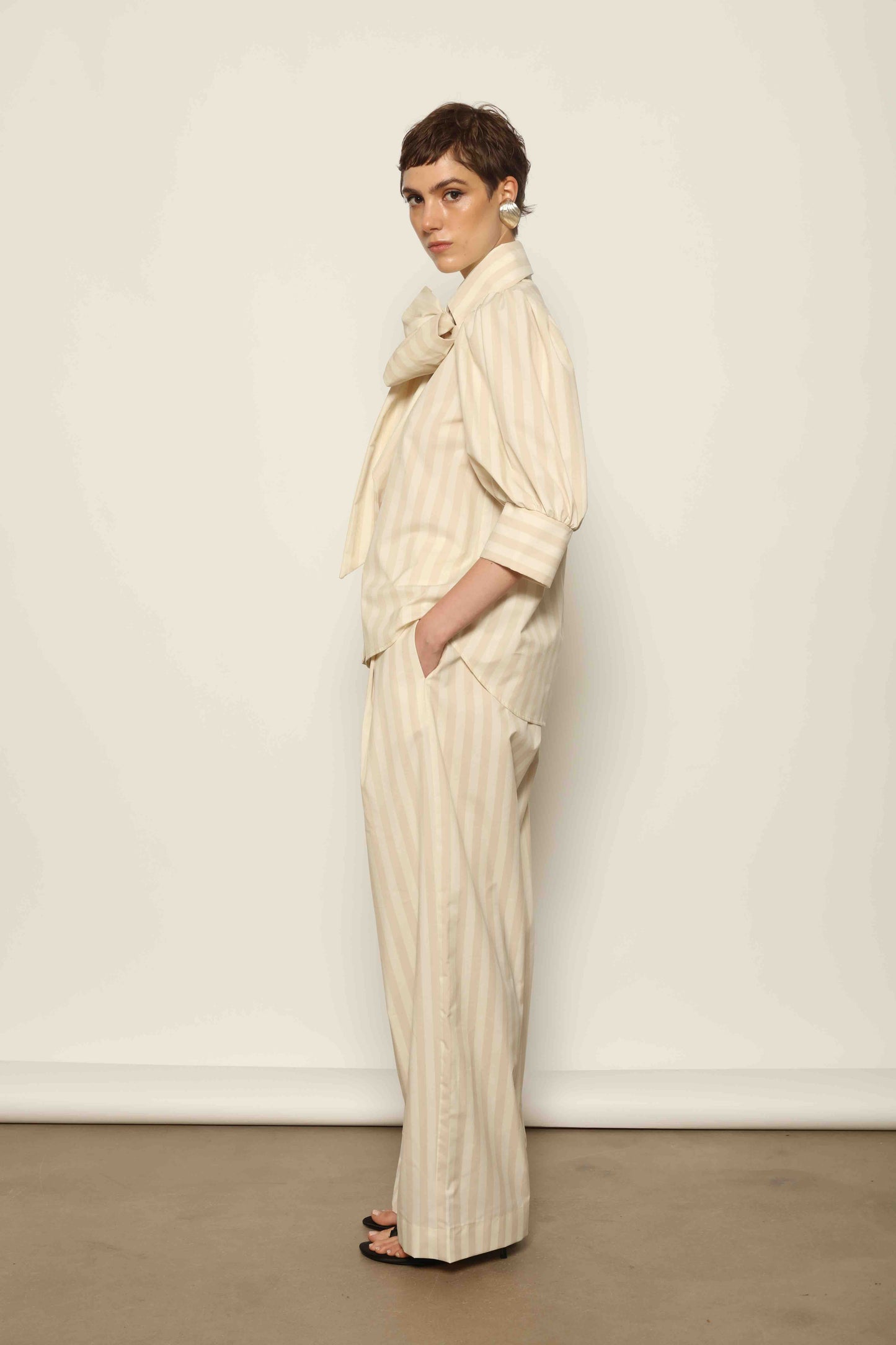 Model wearing the Karin Sand White Macro-Striped Blouse by House of Nomad Stories, featuring a relaxed fit, sand and white macro stripes, classic collar, and soft blend of cotton, merino wool, and cashmere.