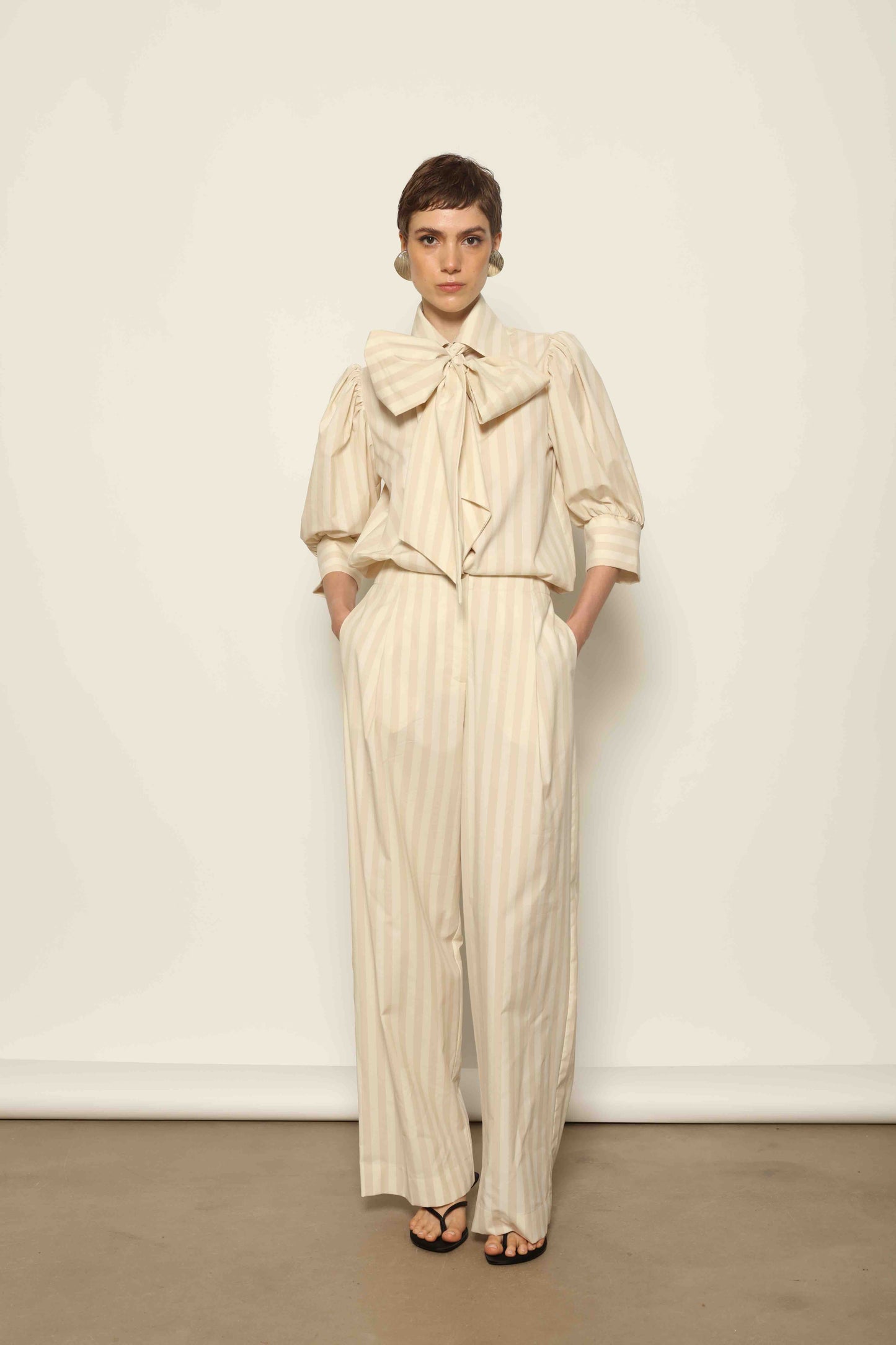 Model wearing the Karin Sand White Macro-Striped Blouse by House of Nomad Stories, featuring a relaxed fit, sand and white macro stripes, classic collar, and soft blend of cotton, merino wool, and cashmere.