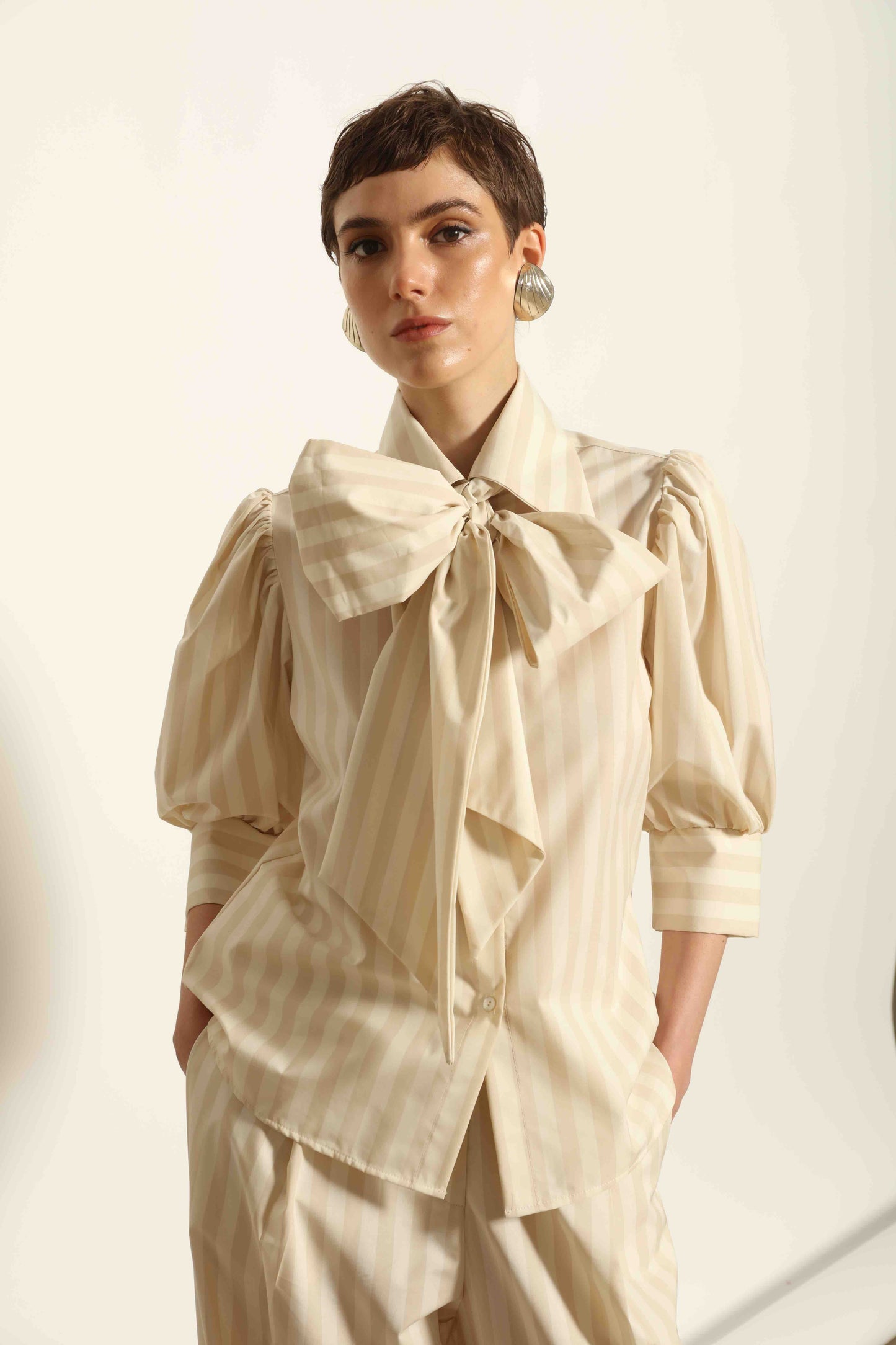 Model wearing the Karin Sand White Macro-Striped Blouse by House of Nomad Stories, featuring a relaxed fit, sand and white macro stripes, classic collar, and soft blend of cotton, merino wool, and cashmere.