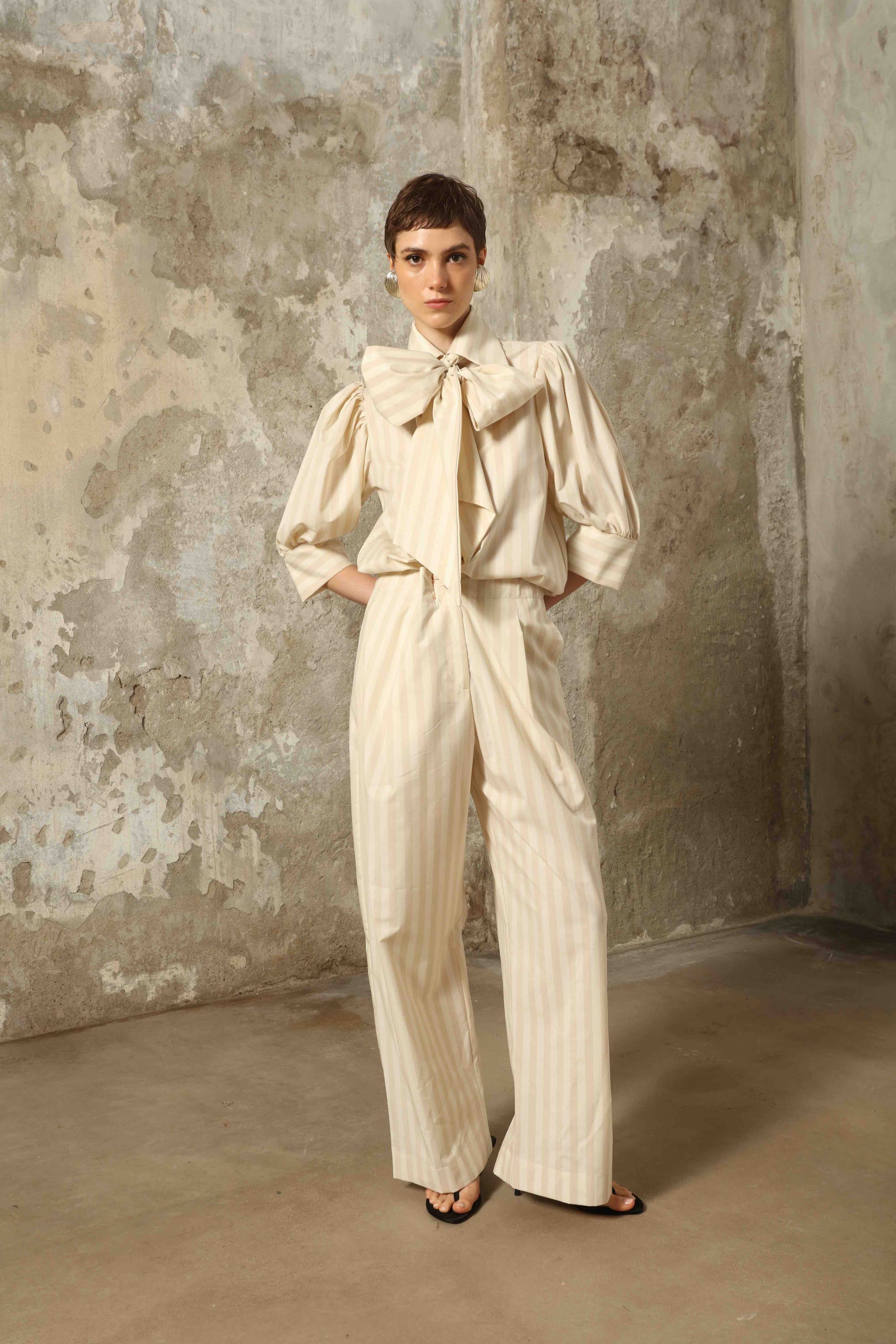 Model wearing the Karin Sand White Macro-Striped Blouse by House of Nomad Stories, featuring a relaxed fit, sand and white macro stripes, classic collar, and soft blend of cotton, merino wool, and cashmere.