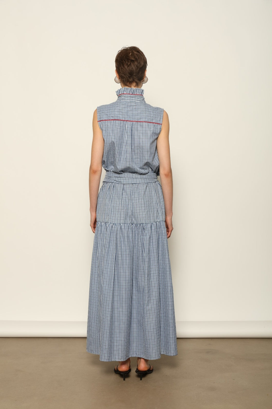 Model wearing the Karin Vichy Blue Dress by House of Woven Stories, featuring 100% organic cotton fabric, blue gingham pattern, sleeveless silhouette, detachable belt, and ruched collar with red piping detail.