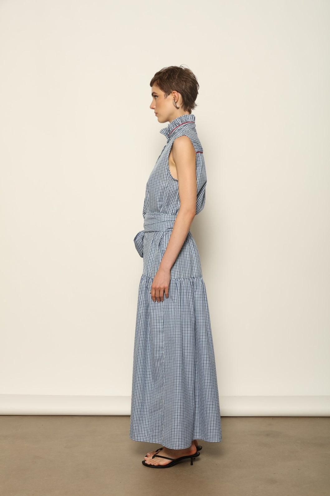 Model wearing the Karin Vichy Blue Dress by House of Woven Stories, featuring 100% organic cotton fabric, blue gingham pattern, sleeveless silhouette, detachable belt, and ruched collar with red piping detail.