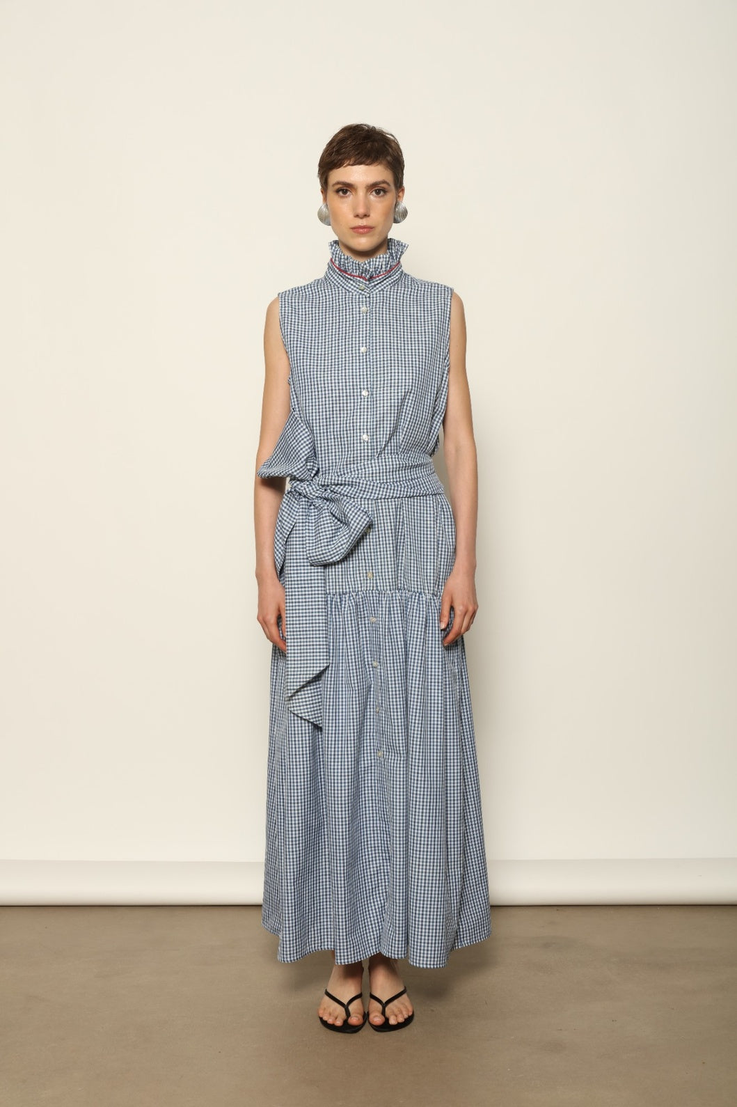 Model wearing the Karin Vichy Blue Dress by House of Woven Stories, featuring 100% organic cotton fabric, blue gingham pattern, sleeveless silhouette, detachable belt, and ruched collar with red piping detail.