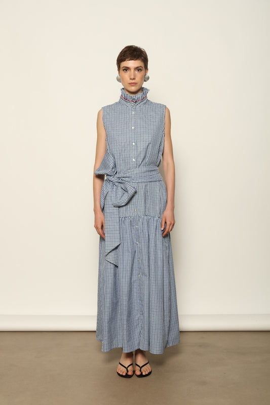 Model wearing the Karin Vichy Blue Dress by House of Woven Stories, featuring 100% organic cotton fabric, blue gingham pattern, sleeveless silhouette, detachable belt, and ruched collar with red piping detail.