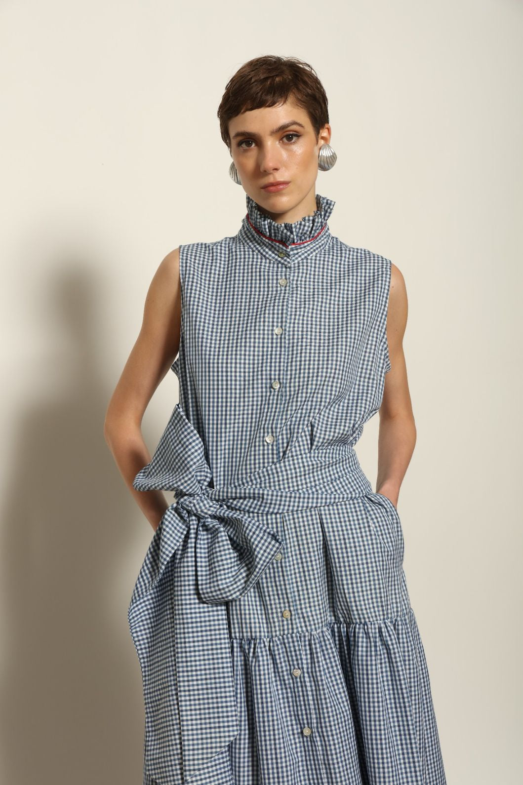 Model wearing the Karin Vichy Blue Dress by House of Woven Stories, featuring 100% organic cotton fabric, blue gingham pattern, sleeveless silhouette, detachable belt, and ruched collar with red piping detail.