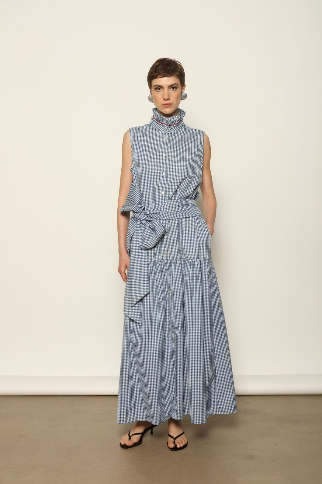 Model wearing the Karin Vichy Blue Dress by House of Woven Stories, featuring 100% organic cotton fabric, blue gingham pattern, sleeveless silhouette, detachable belt, and ruched collar with red piping detail.