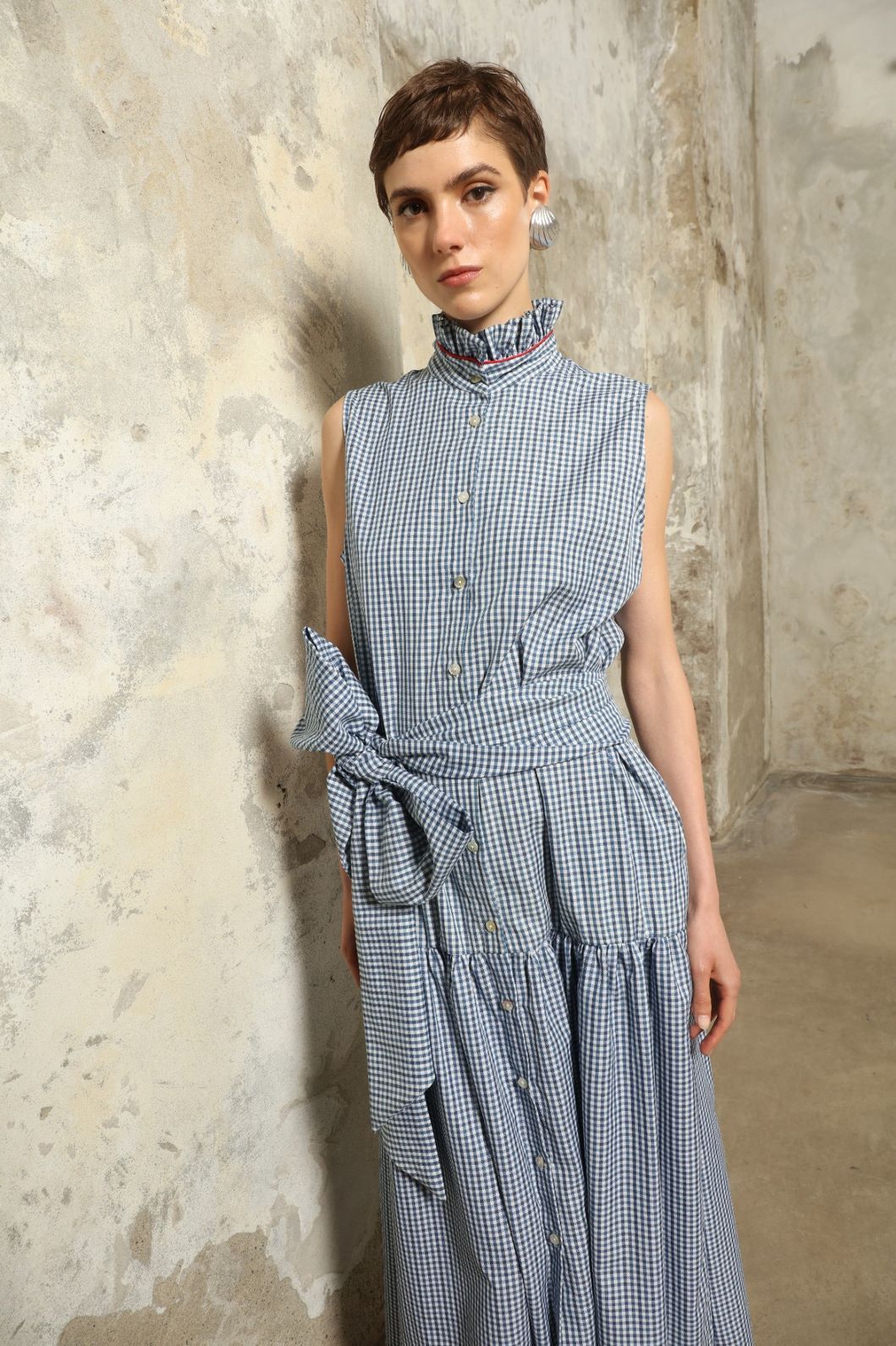 Model wearing the Karin Vichy Blue Dress by House of Woven Stories, featuring 100% organic cotton fabric, blue gingham pattern, sleeveless silhouette, detachable belt, and ruched collar with red piping detail.