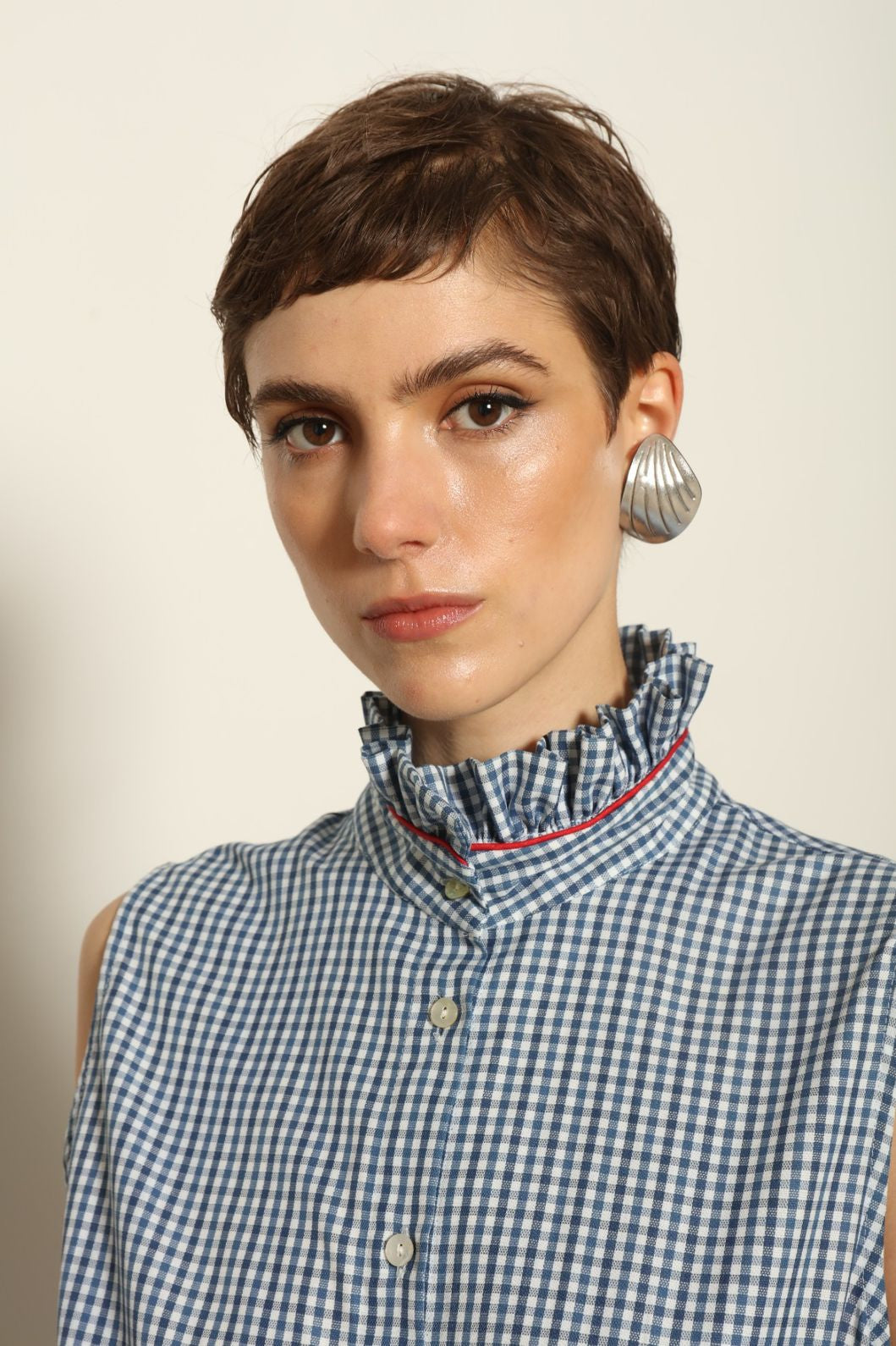 Model wearing the Karin Vichy Blue Dress by House of Woven Stories, featuring 100% organic cotton fabric, blue gingham pattern, sleeveless silhouette, detachable belt, and ruched collar with red piping detail.