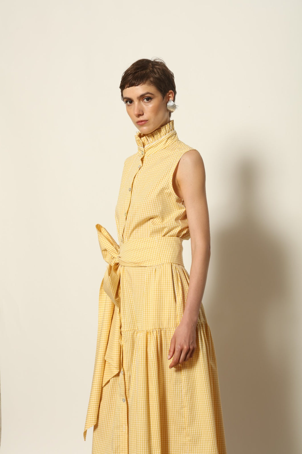 Model wearing the Karin Vichy Butter Yellow Dress by House of Woven Stories, featuring 100% organic cotton fabric, butter yellow gingham pattern, sleeveless silhouette, signature ruffled collar, and white piping details.