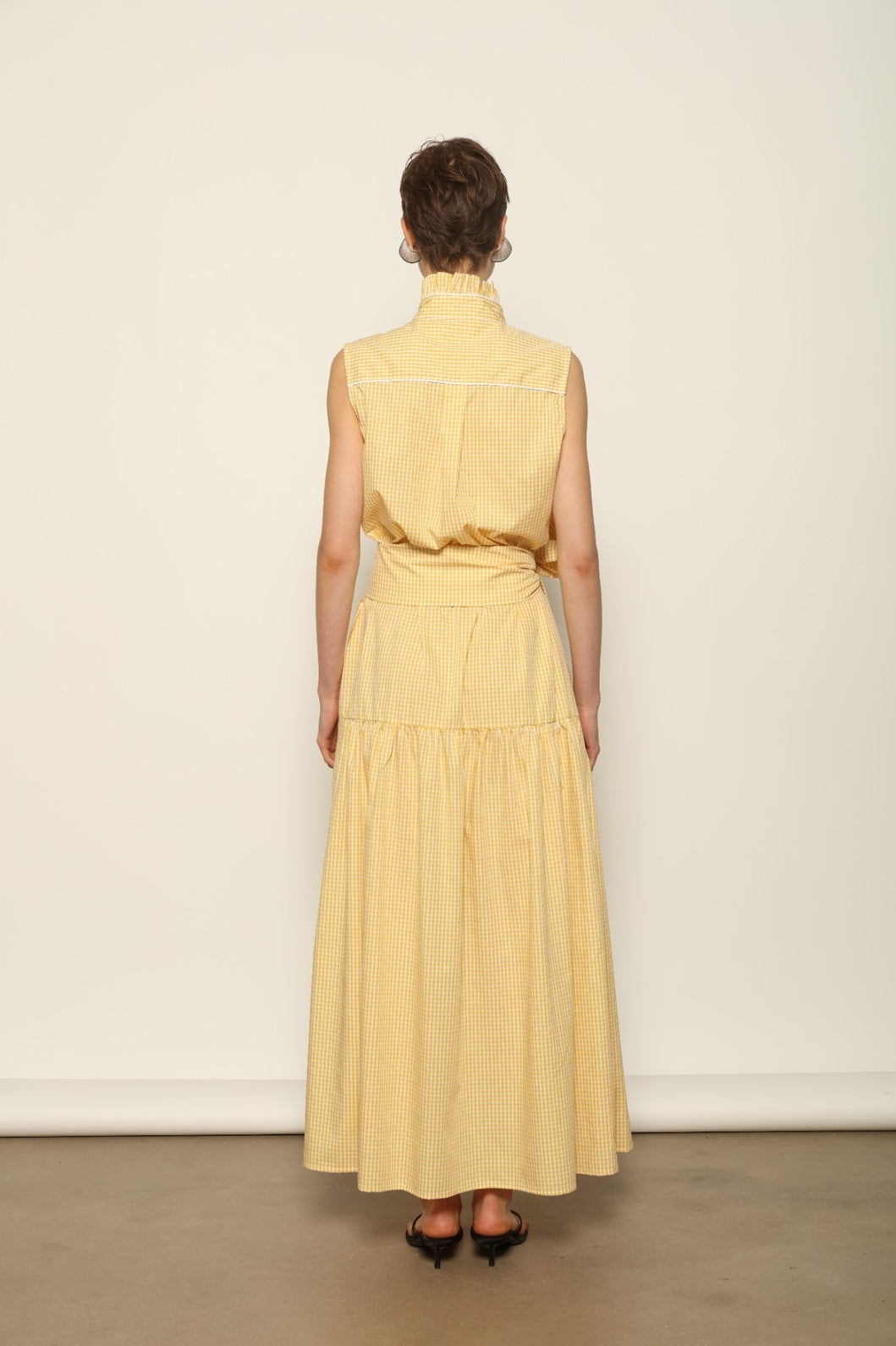 Model wearing the Karin Vichy Butter Yellow Dress by House of Woven Stories, featuring 100% organic cotton fabric, butter yellow gingham pattern, sleeveless silhouette, signature ruffled collar, and white piping details.