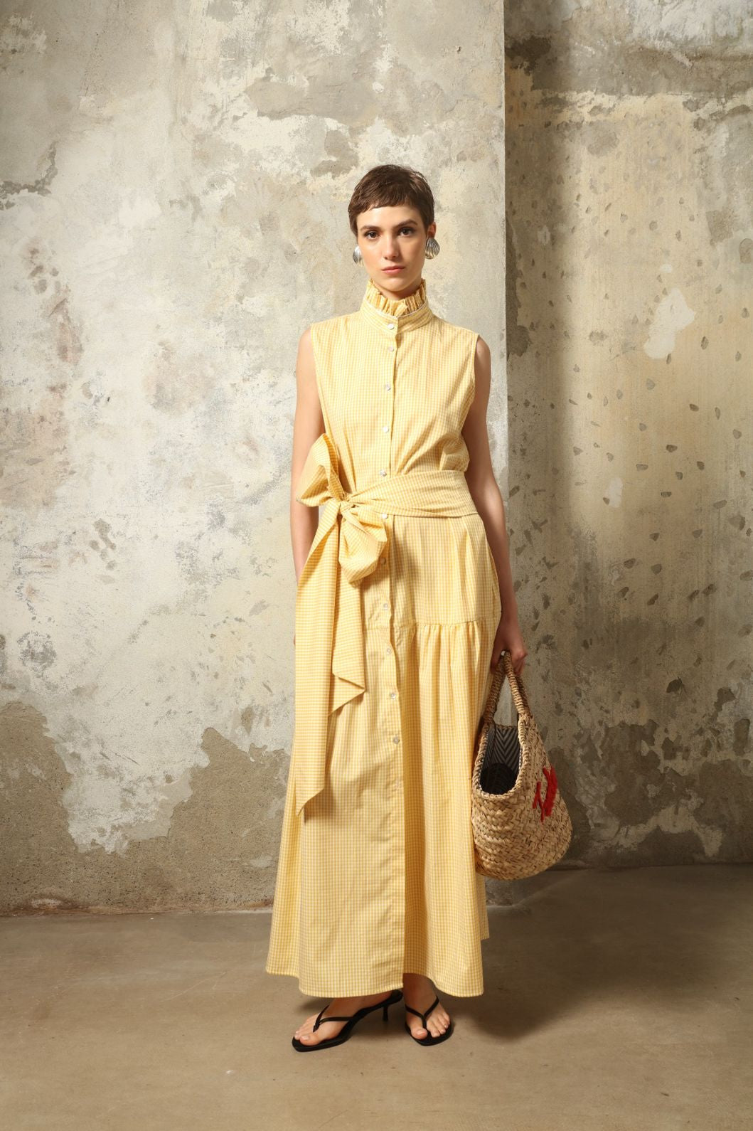 Model wearing the Karin Vichy Butter Yellow Dress by House of Woven Stories, featuring 100% organic cotton fabric, butter yellow gingham pattern, sleeveless silhouette, signature ruffled collar, and white piping details.