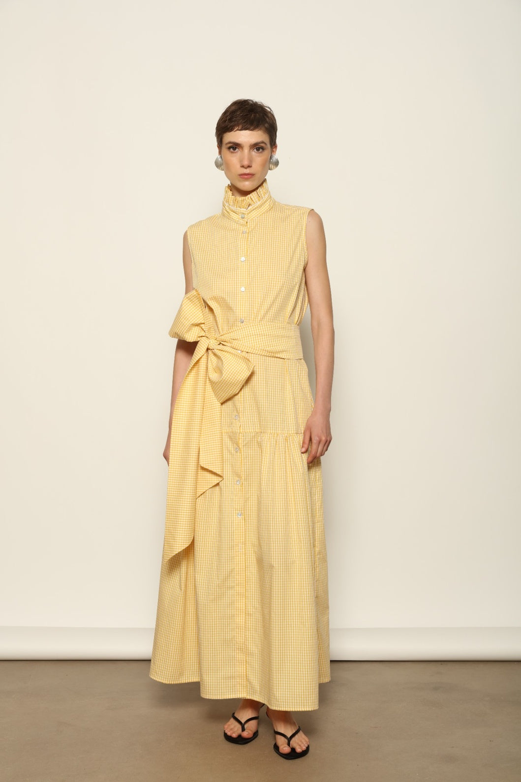 Model wearing the Karin Vichy Butter Yellow Dress by House of Woven Stories, featuring 100% organic cotton fabric, butter yellow gingham pattern, sleeveless silhouette, signature ruffled collar, and white piping details.