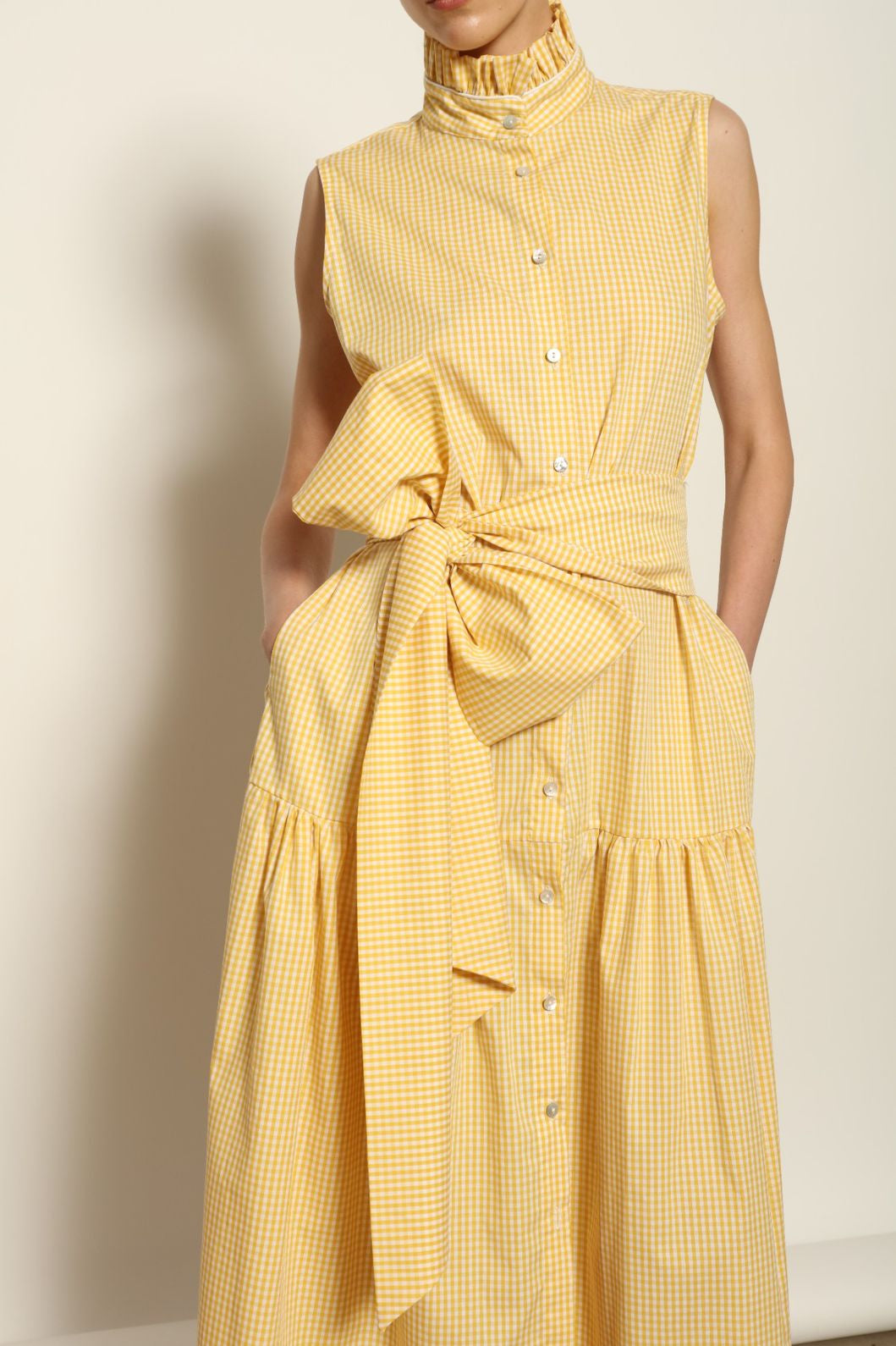 Model wearing the Karin Vichy Butter Yellow Dress by House of Woven Stories, featuring 100% organic cotton fabric, butter yellow gingham pattern, sleeveless silhouette, signature ruffled collar, and white piping details.