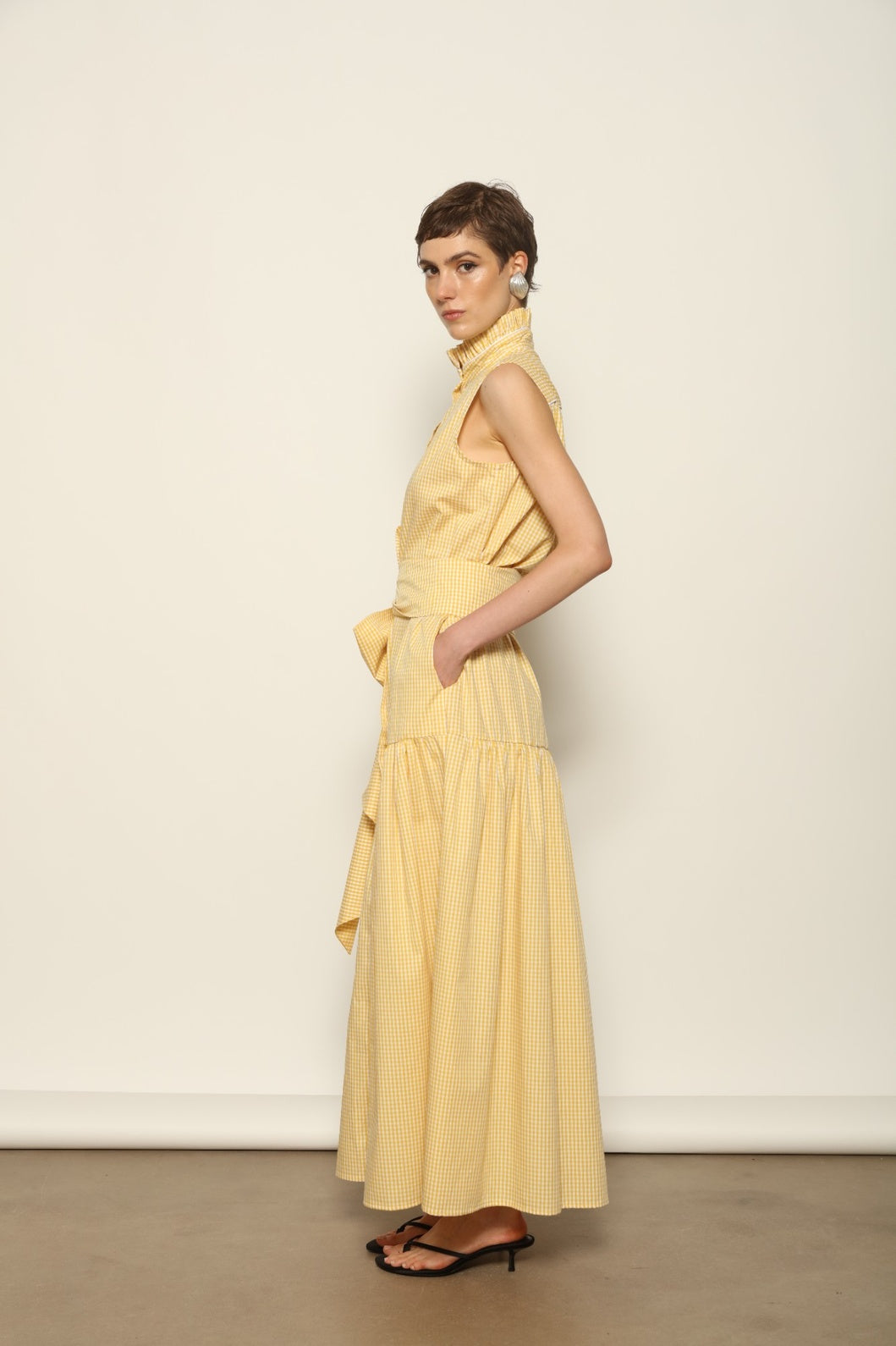 Model wearing the Karin Vichy Butter Yellow Dress by House of Woven Stories, featuring 100% organic cotton fabric, butter yellow gingham pattern, sleeveless silhouette, signature ruffled collar, and white piping details.