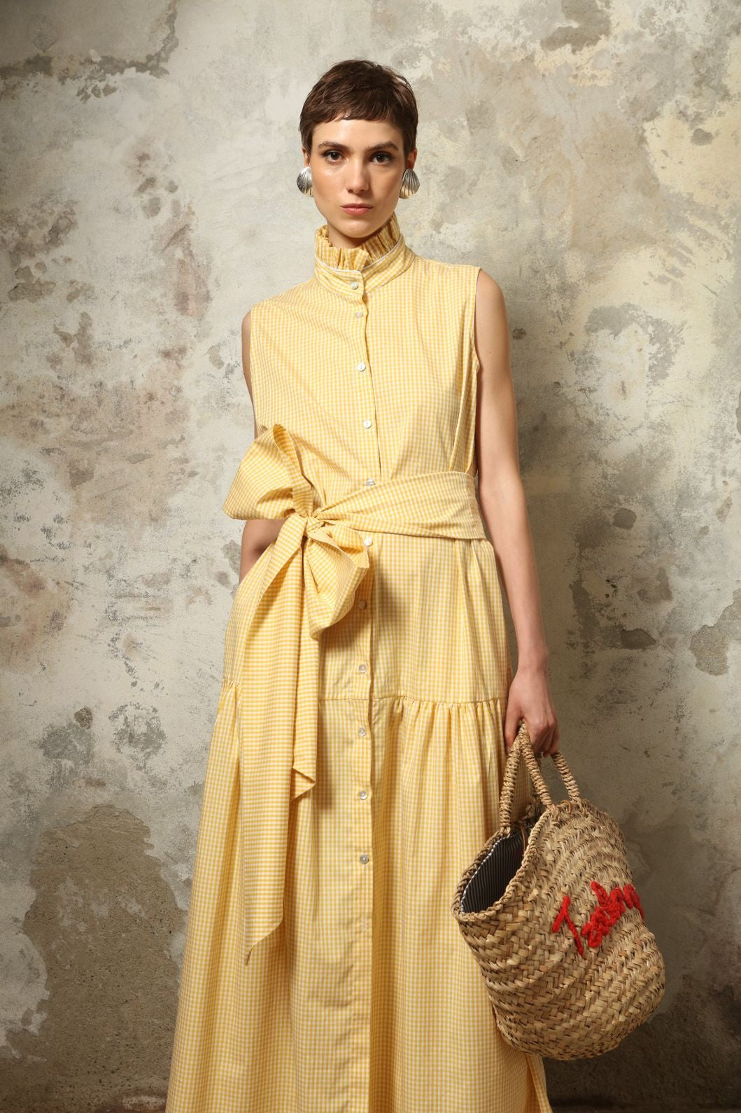 Model wearing the Karin Vichy Butter Yellow Dress by House of Woven Stories, featuring 100% organic cotton fabric, butter yellow gingham pattern, sleeveless silhouette, signature ruffled collar, and white piping details.