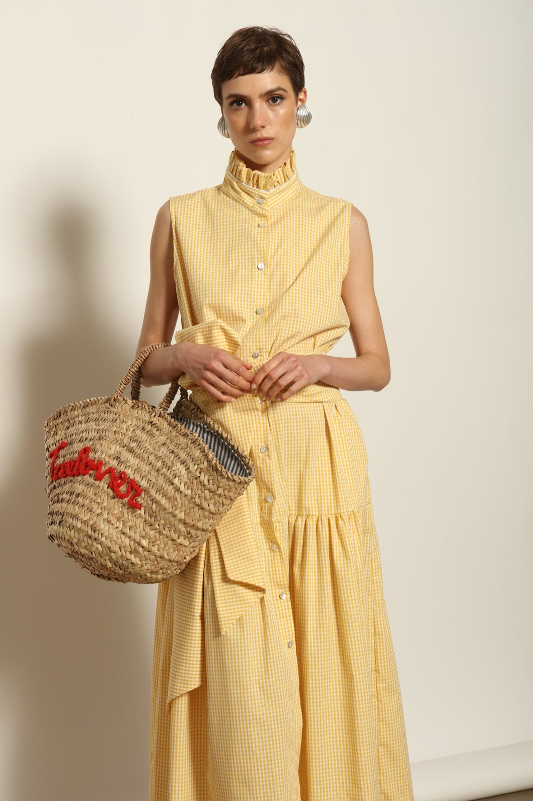 Model wearing the Karin Vichy Butter Yellow Dress by House of Woven Stories, featuring 100% organic cotton fabric, butter yellow gingham pattern, sleeveless silhouette, signature ruffled collar, and white piping details.