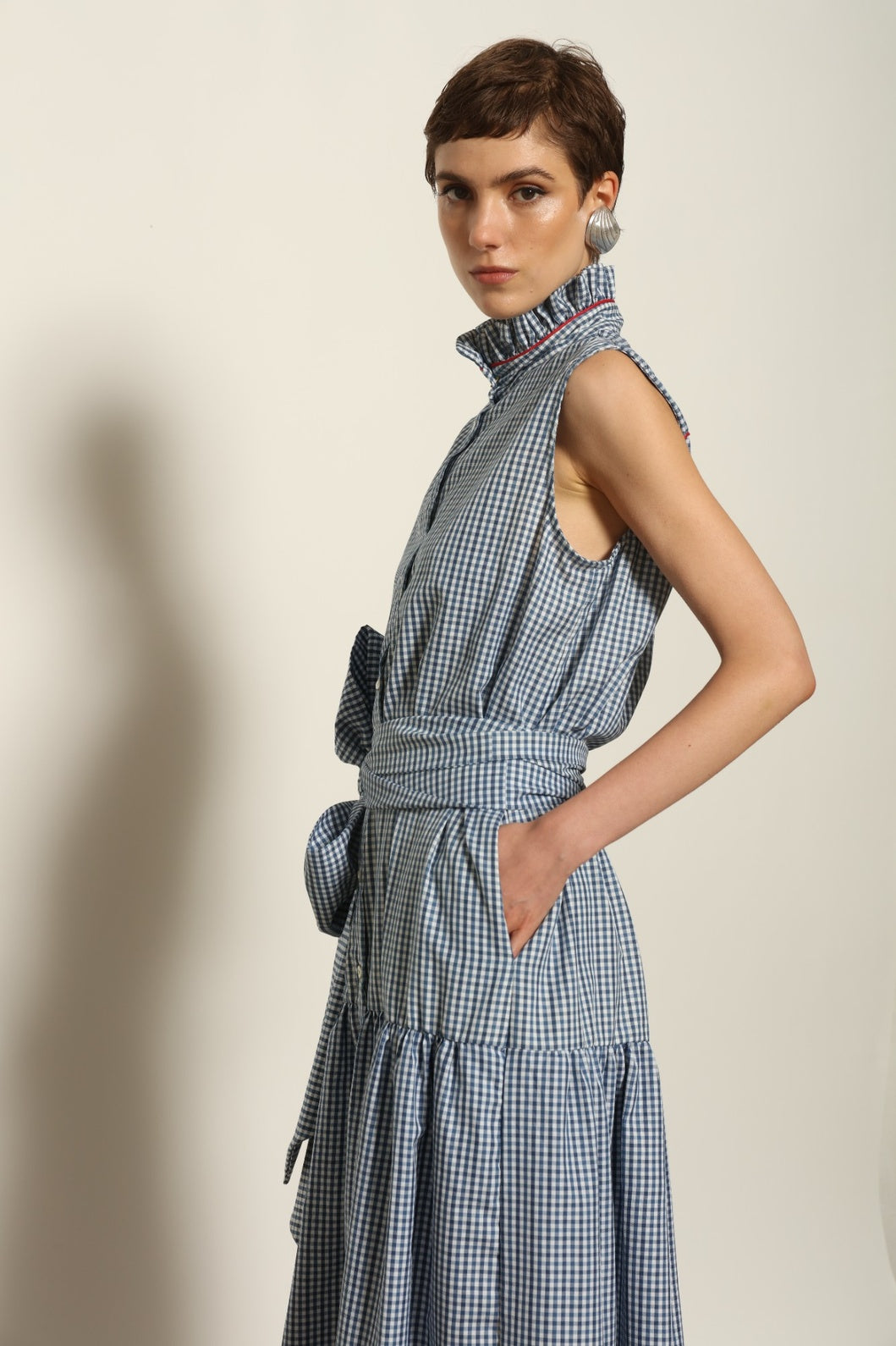 Model wearing the Karin Vichy Blue Dress by House of Woven Stories, featuring 100% organic cotton fabric, blue gingham pattern, sleeveless silhouette, detachable belt, and ruched collar with red piping detail.