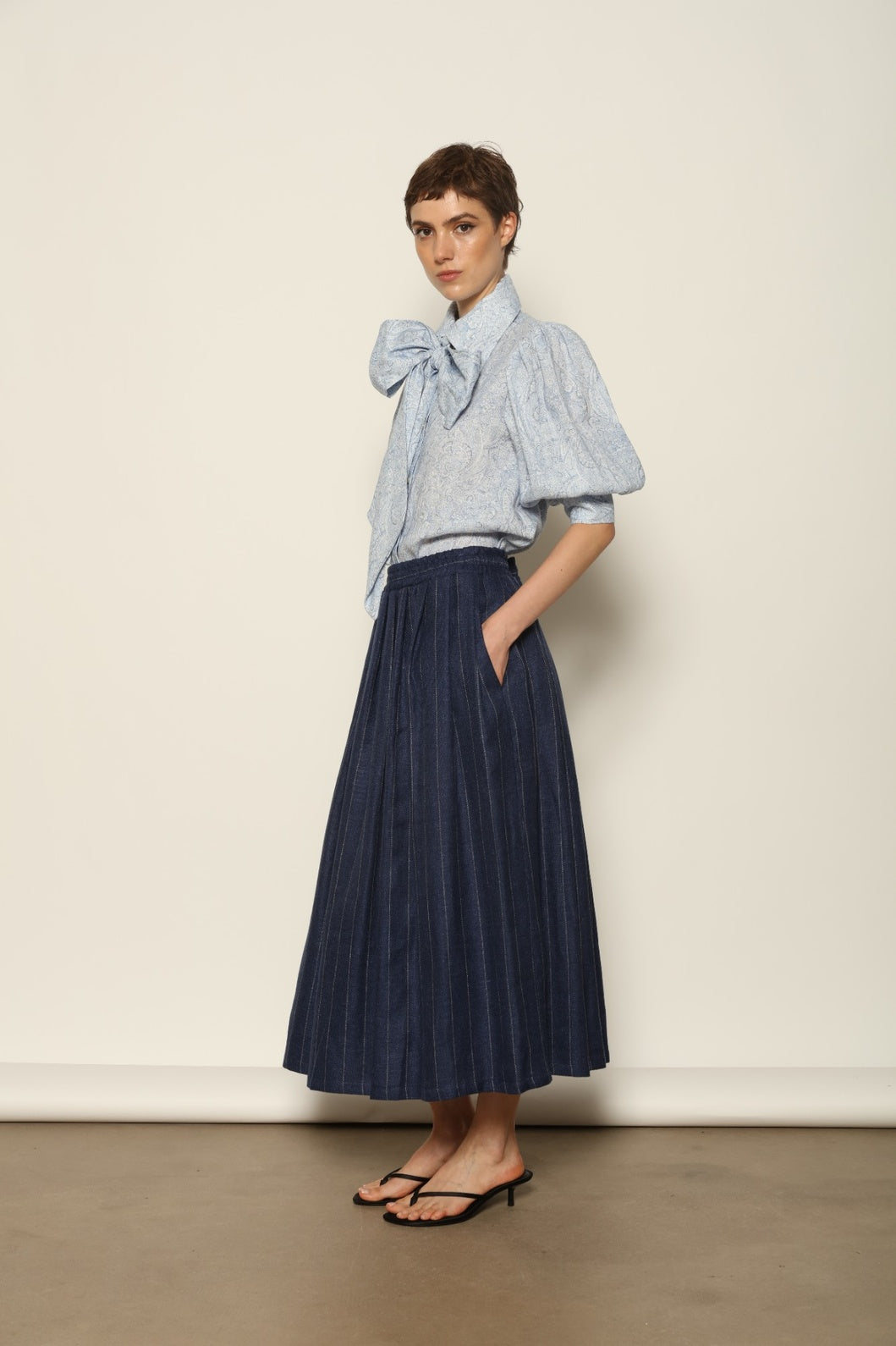 Model wearing the Karin White Blue Paisley Blouse by House of Nomad Stories, featuring 100% linen fabric, blue and white paisley print, relaxed short sleeves, and a classic collar with tie detail.