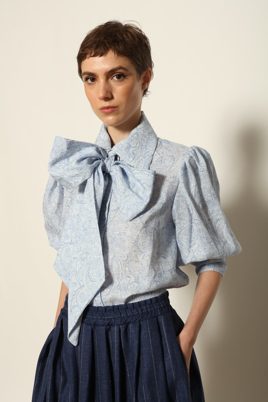 Model wearing the Karin White Blue Paisley Blouse by House of Nomad Stories, featuring 100% linen fabric, blue and white paisley print, relaxed short sleeves, and a classic collar with tie detail.