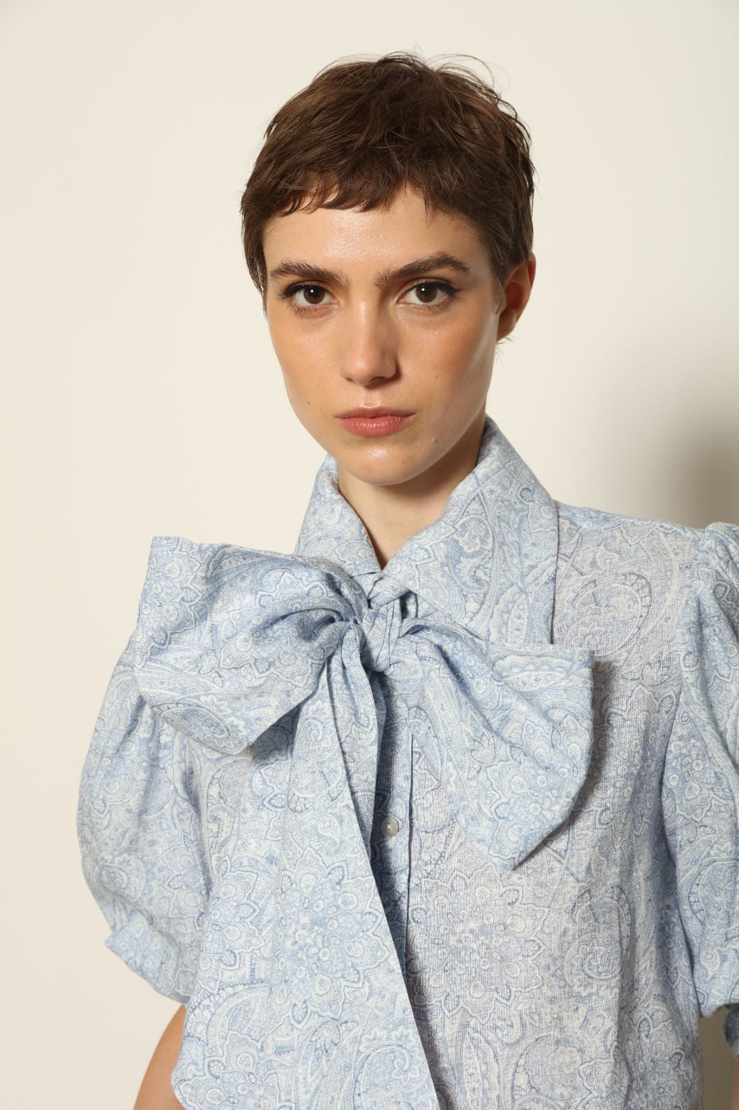 Model wearing the Karin White Blue Paisley Blouse by House of Nomad Stories, featuring 100% linen fabric, blue and white paisley print, relaxed short sleeves, and a classic collar with tie detail.