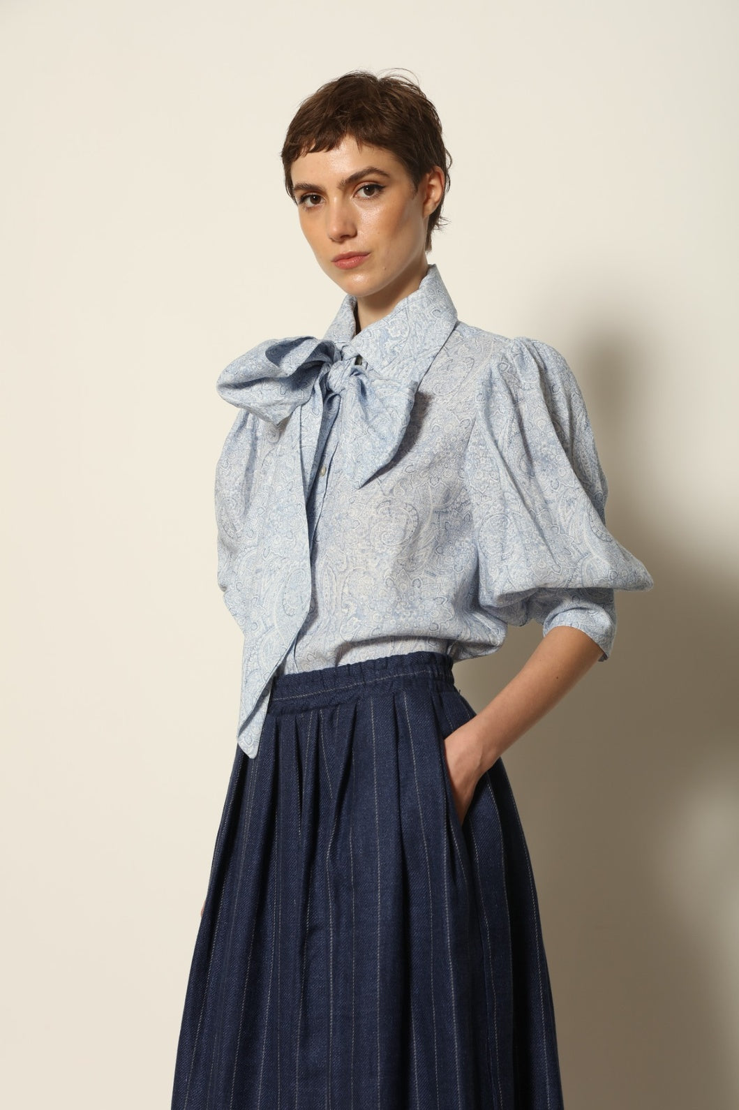 Model wearing the Karin White Blue Paisley Blouse by House of Nomad Stories, featuring 100% linen fabric, blue and white paisley print, relaxed short sleeves, and a classic collar with tie detail.