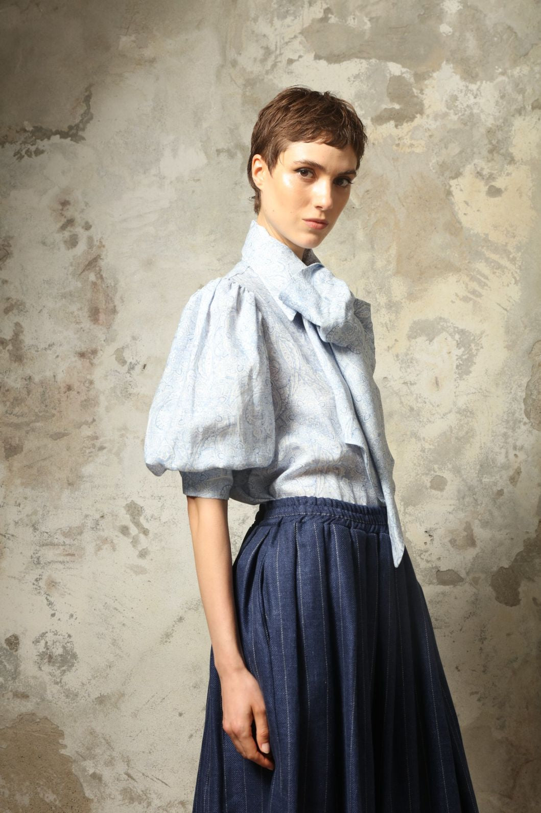 Model wearing the Karin White Blue Paisley Blouse by House of Nomad Stories, featuring 100% linen fabric, blue and white paisley print, relaxed short sleeves, and a classic collar with tie detail.