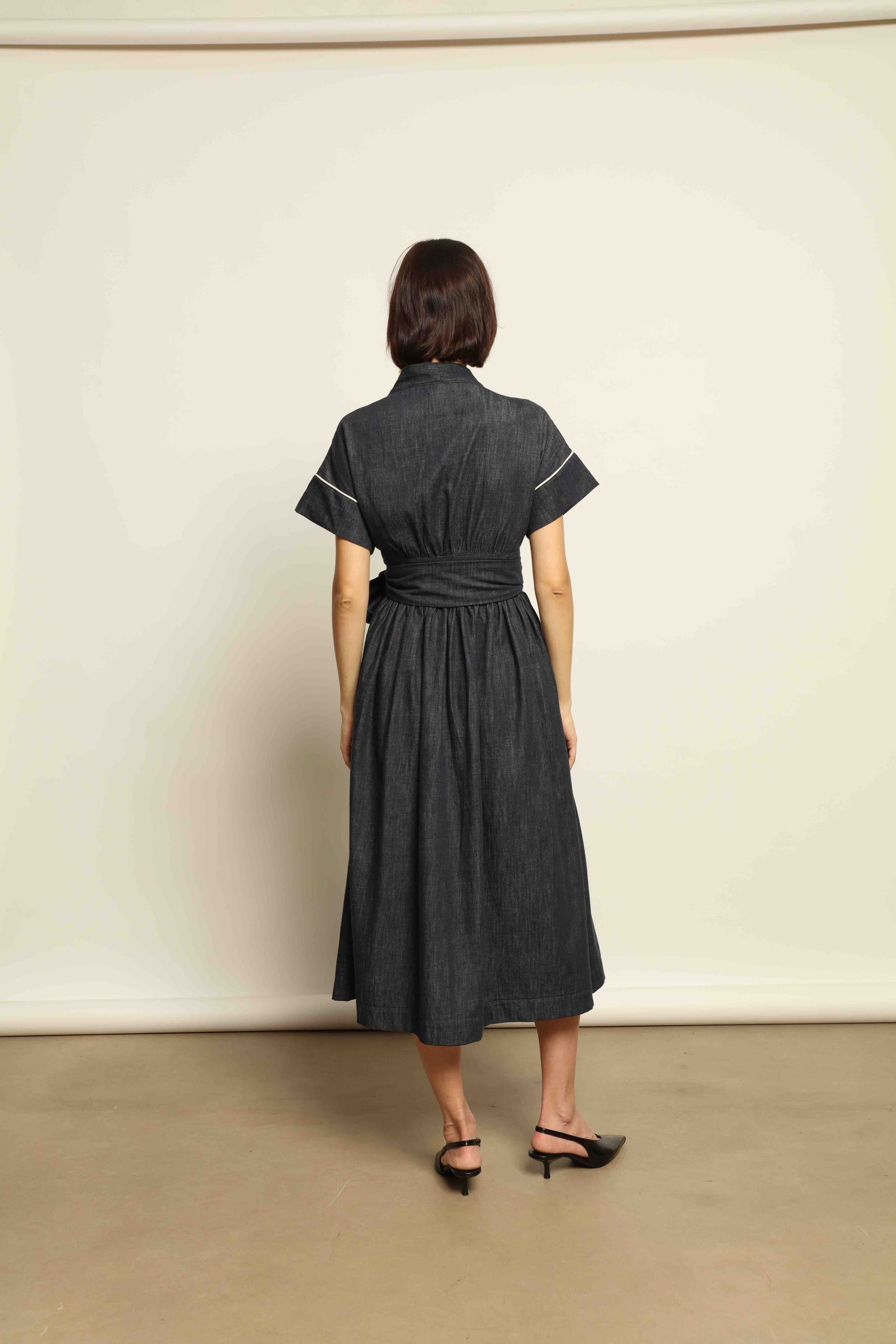 Model is wearing the Saint Remy wrap dress by House of Woven Stories in 100% cotton, featuring a denim-blue color with white pinstripes, drop shoulders, short sleeves, deep V-neckline, and self-tie belt, handmade in Italy.
