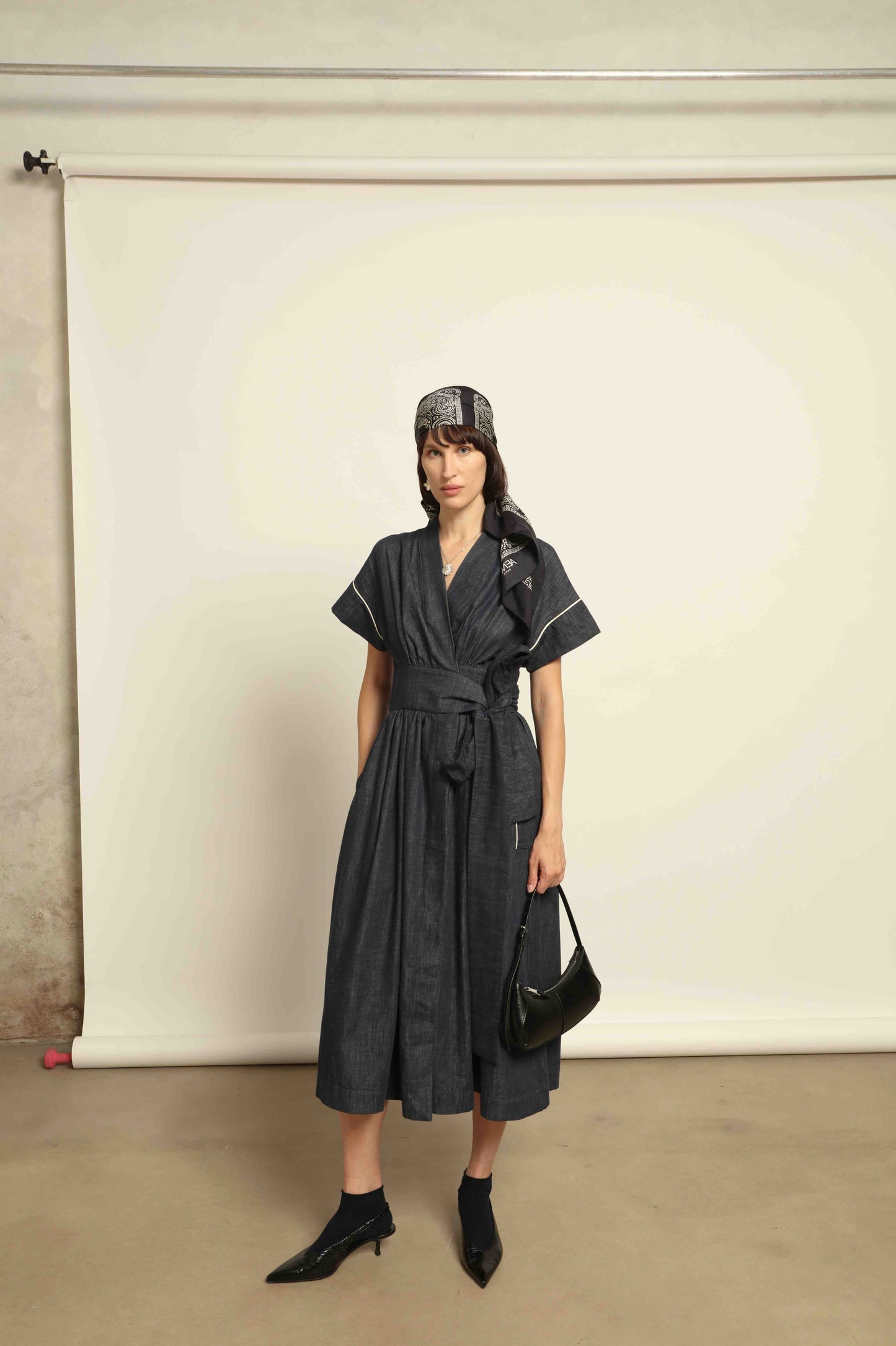 Model is wearing the Saint Remy wrap dress by House of Woven Stories in 100% cotton, featuring a denim-blue color with white pinstripes, drop shoulders, short sleeves, deep V-neckline, and self-tie belt, handmade in Italy.