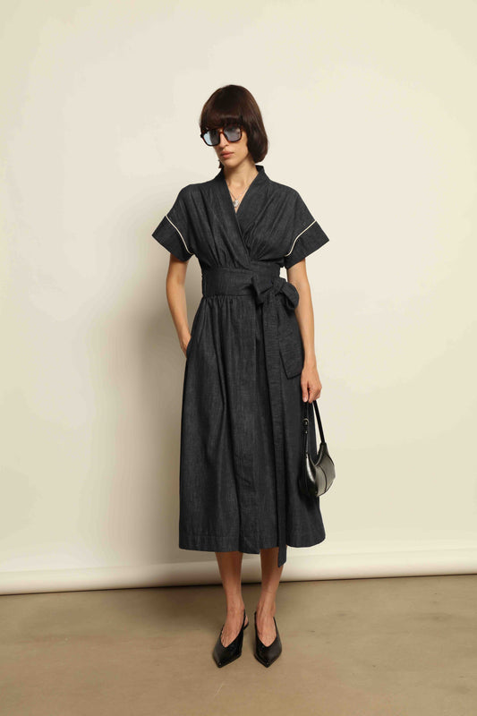 Model is wearing the Saint Remy wrap dress by House of Woven Stories in 100% cotton, featuring a denim-blue color with white pinstripes, drop shoulders, short sleeves, deep V-neckline, and self-tie belt, handmade in Italy.