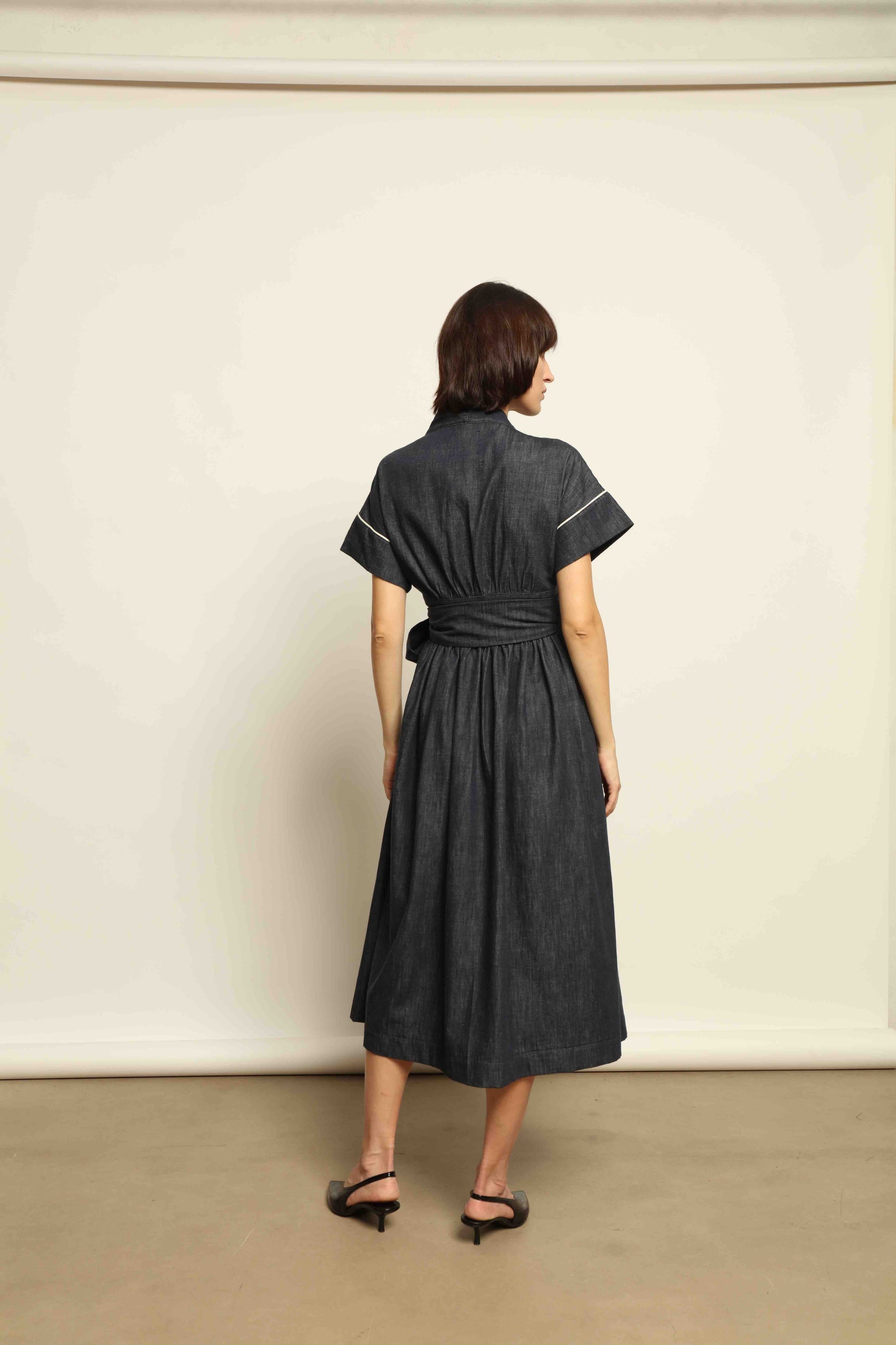 Model is wearing the Saint Remy wrap dress by House of Woven Stories in 100% cotton, featuring a denim-blue color with white pinstripes, drop shoulders, short sleeves, deep V-neckline, and self-tie belt, handmade in Italy.