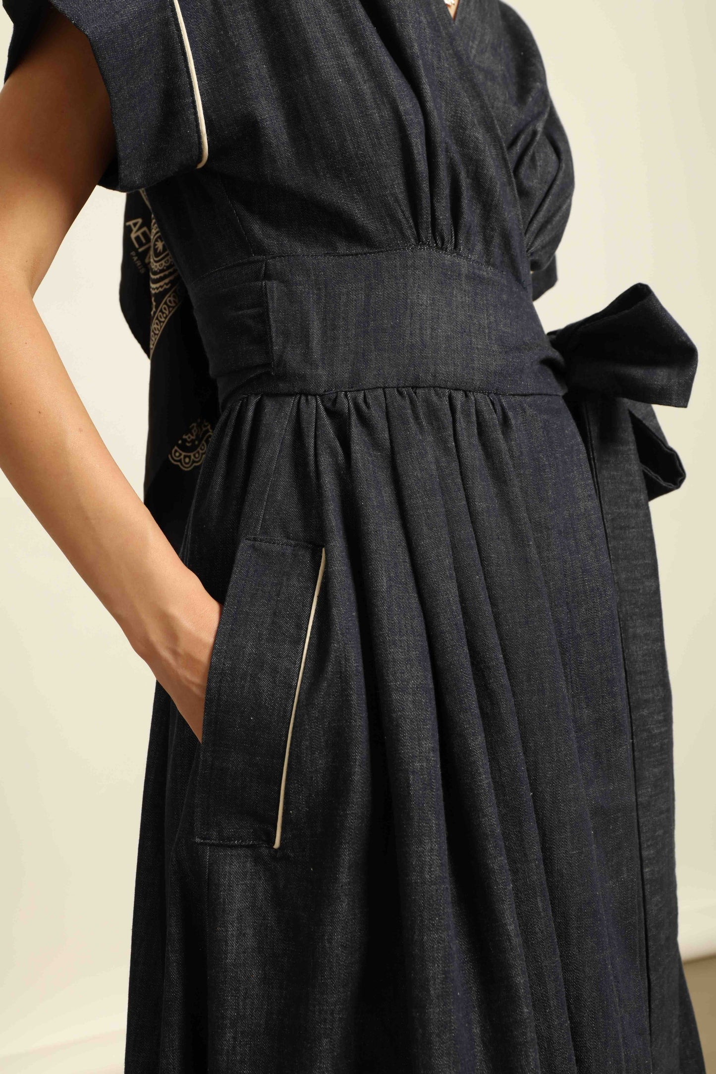 Model is wearing the Saint Remy wrap dress by House of Woven Stories in 100% cotton, featuring a denim-blue color with white pinstripes, drop shoulders, short sleeves, deep V-neckline, and self-tie belt, handmade in Italy.