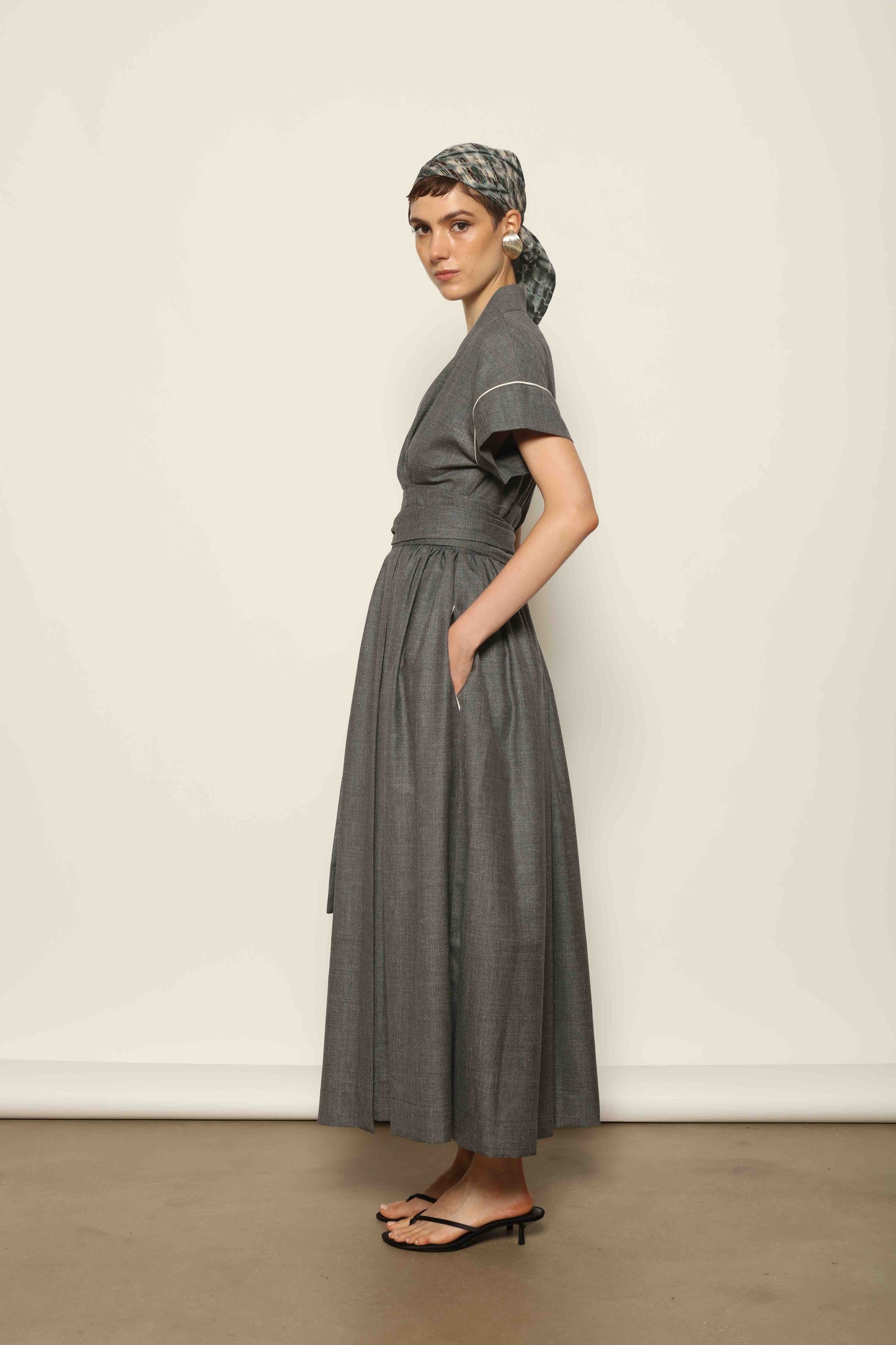 Model wearing the Saint Remy Grey Off White Midi Dress by House of Woven Stories, featuring a wrap silhouette, adjustable belt, half sleeves, and subtle white piping detail in a fluid elegant fabric.