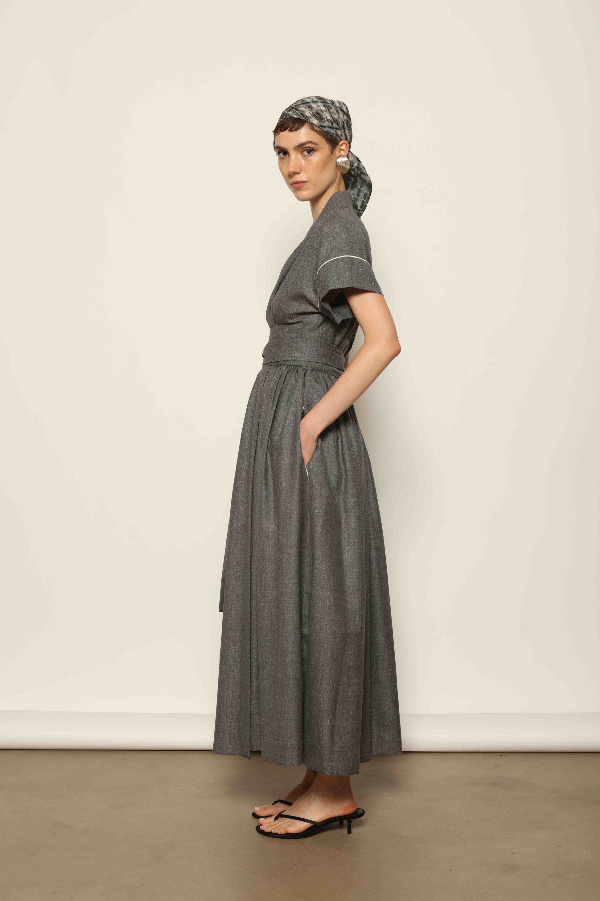 Model wearing the Saint Remy Grey Off White Midi Dress by House of Woven Stories, featuring a wrap silhouette, adjustable belt, half sleeves, and subtle white piping detail in a fluid elegant fabric.