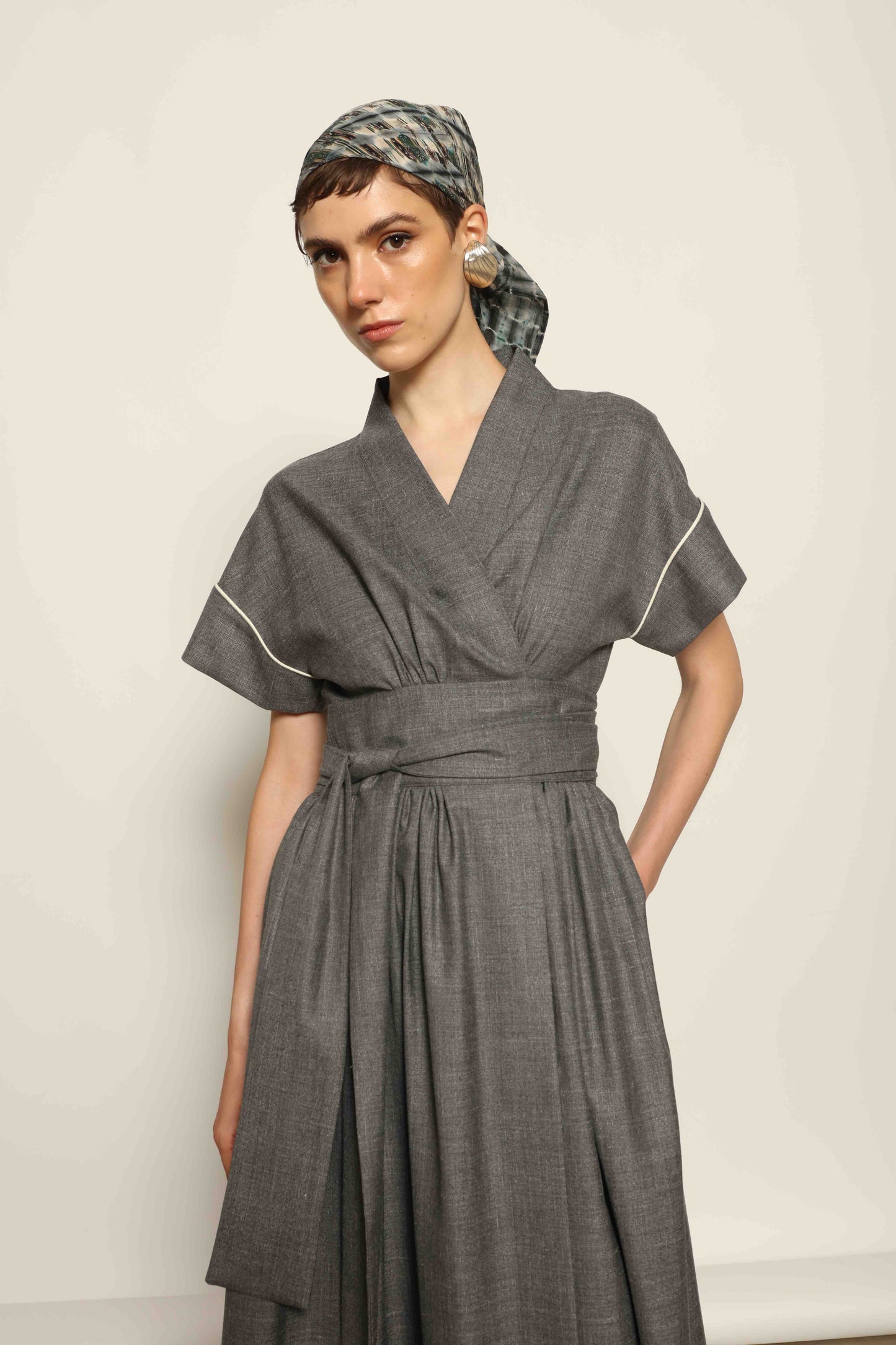 Model wearing the Saint Remy Grey Off White Midi Dress by House of Woven Stories, featuring a wrap silhouette, adjustable belt, half sleeves, and subtle white piping detail in a fluid elegant fabric.