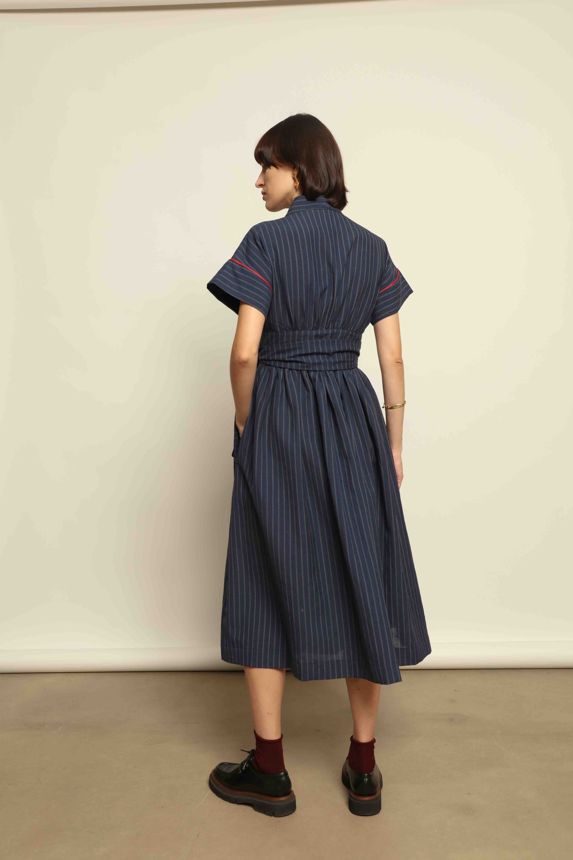 Model is wearing the Saint Remy wrap dress by House of Woven Stories in a blue and white pinstripe, made from a breathable wool-linen blend, featuring drop shoulders, short sleeves, deep V-neckline, and a matching fabric belt, handmade in Italy.