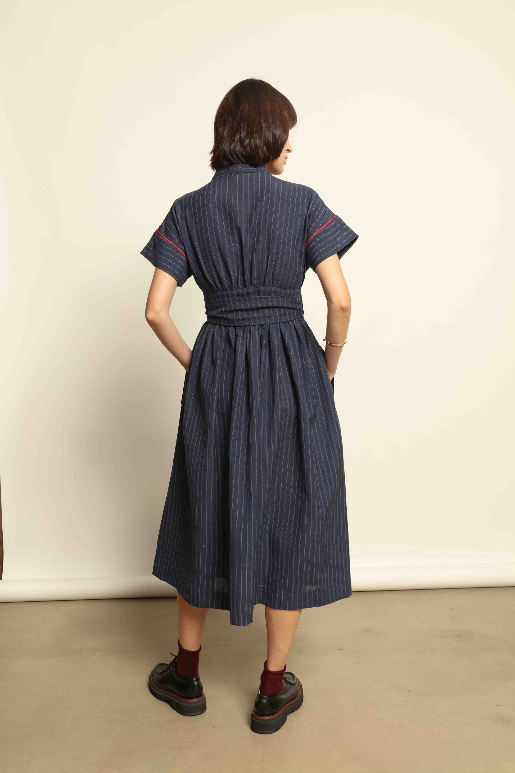 Model is wearing the Saint Remy wrap dress by House of Woven Stories in a blue and white pinstripe, made from a breathable wool-linen blend, featuring drop shoulders, short sleeves, deep V-neckline, and a matching fabric belt, handmade in Italy.