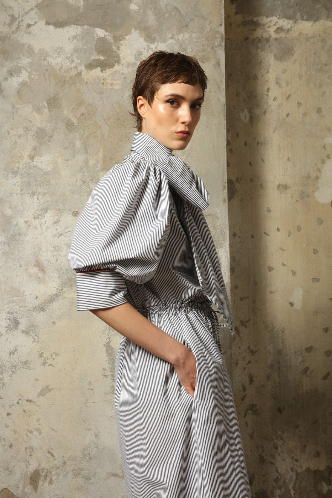 Model wearing the Karin White Navy Striped Blouse by House of Woven Stories, featuring organic cotton fabric, white and navy stripes, red piping detail, short sleeves, and a relaxed elegant silhouette.