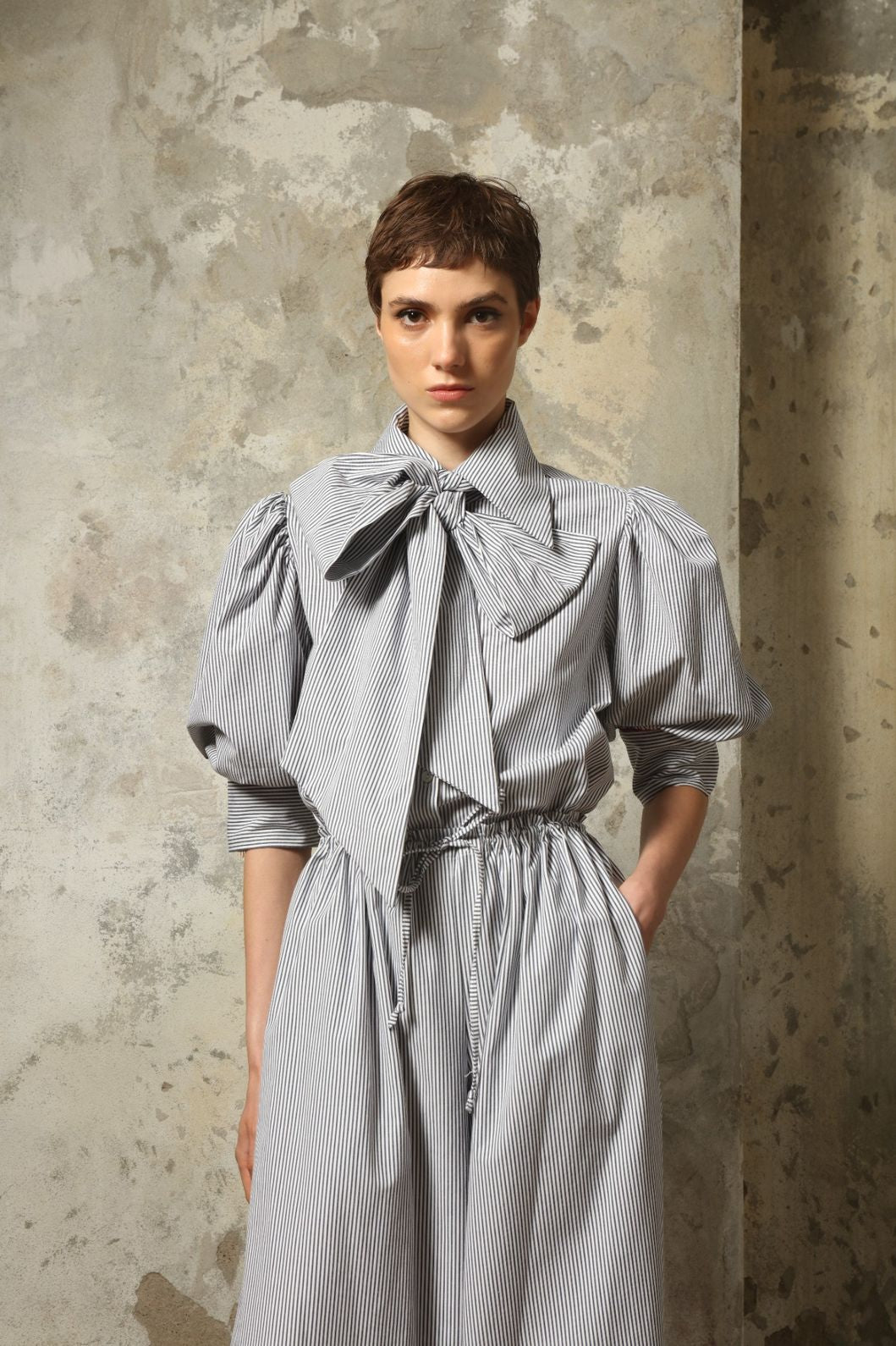 Model wearing the Karin White Navy Striped Blouse by House of Woven Stories, featuring organic cotton fabric, white and navy stripes, red piping detail, short sleeves, and a relaxed elegant silhouette.