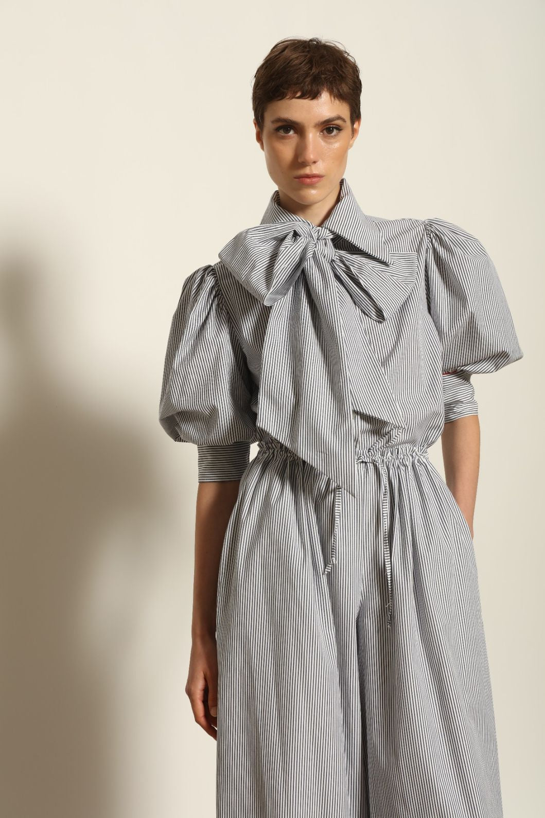 Model wearing the Karin White Navy Striped Blouse by House of Woven Stories, featuring organic cotton fabric, white and navy stripes, red piping detail, short sleeves, and a relaxed elegant silhouette.