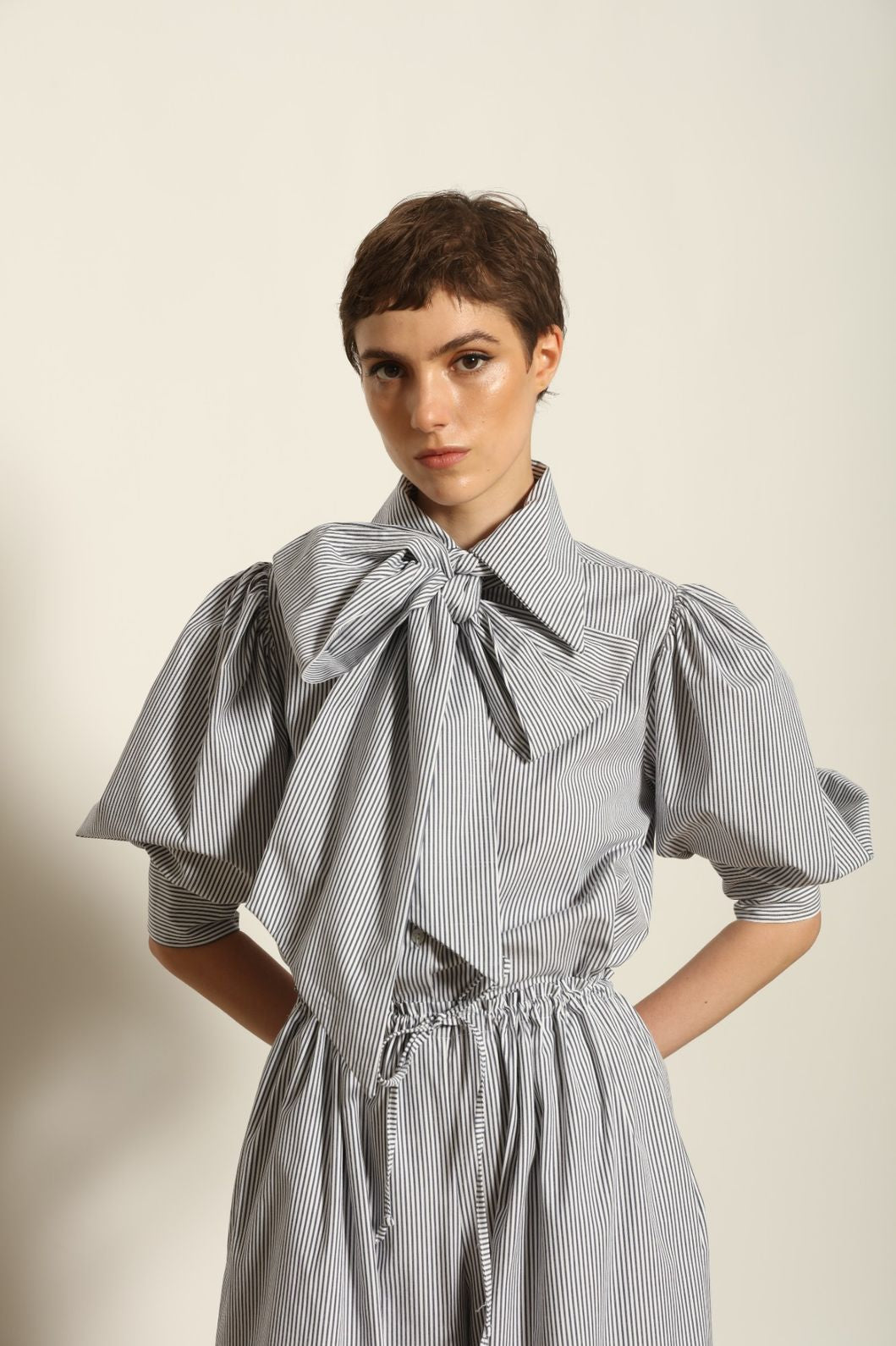 Model wearing the Karin White Navy Striped Blouse by House of Woven Stories, featuring organic cotton fabric, white and navy stripes, red piping detail, short sleeves, and a relaxed elegant silhouette.