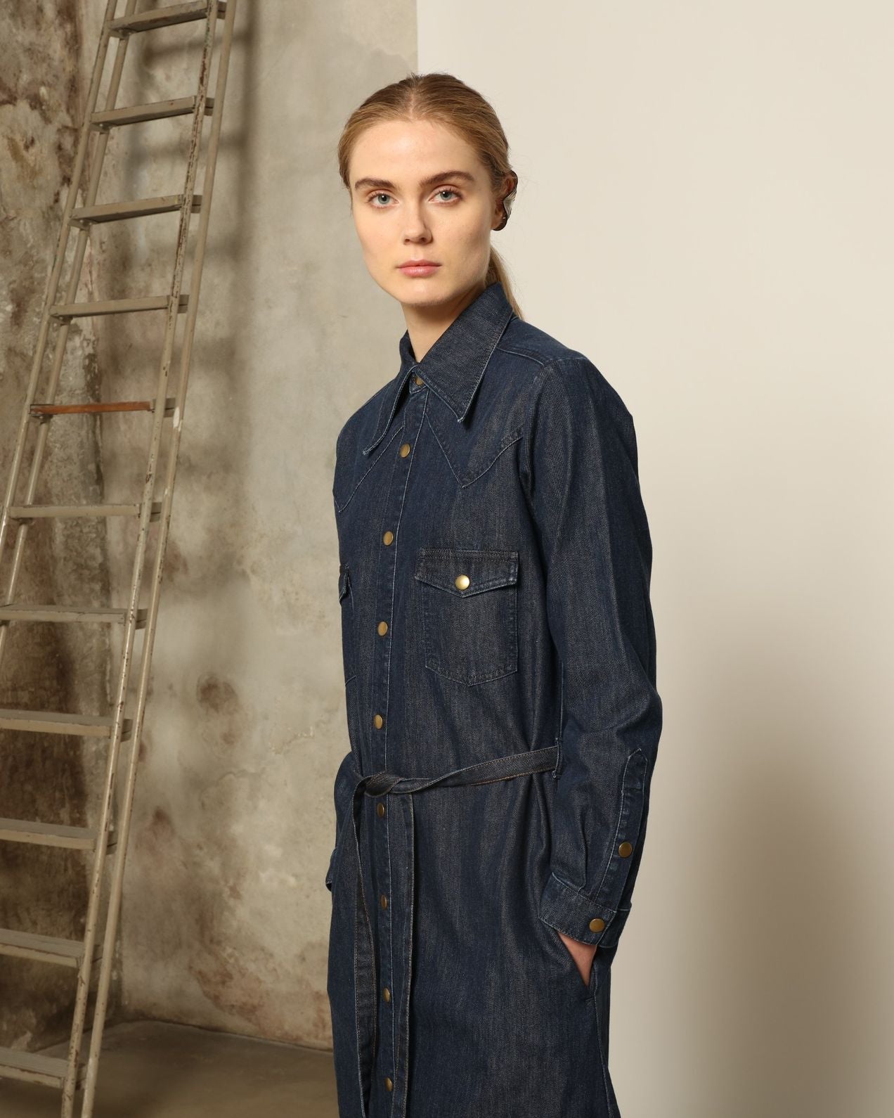 Model is wearing La Robe Star in Bleu D’été by Happy Haus, a denim shirt dress with relaxed fit, chest pockets, matching belt, and golden button detail, combining timeless style with versatile year-round wear.