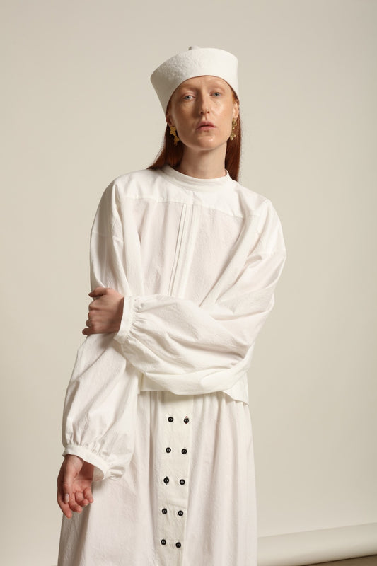 Model is wearing the Les Vacances d’Irina white cotton blouse in 100% cotton with voluminous sleeves and pleated front, from the House of Nomad Stories selection.