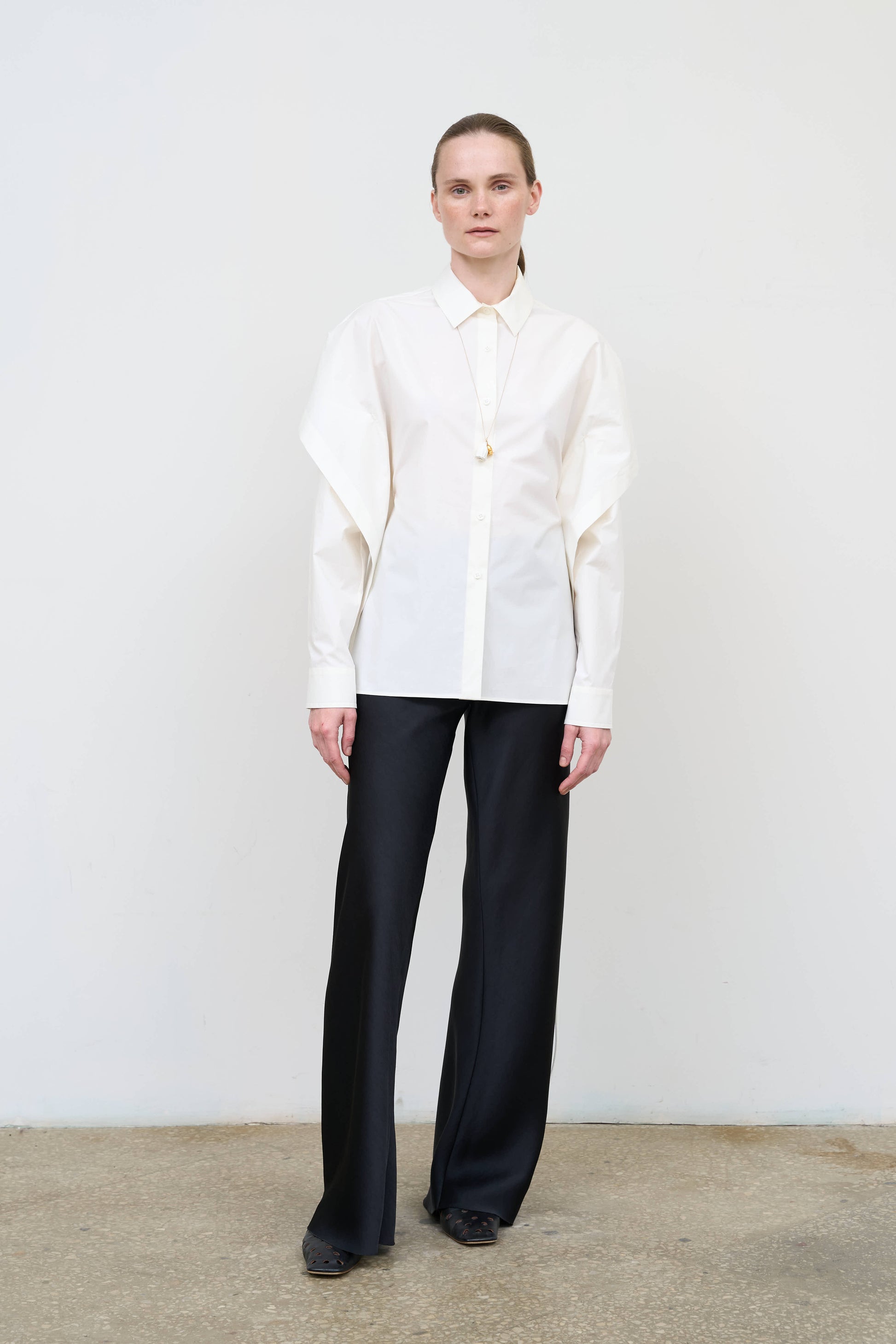 Model is wearing a fitted white cotton shirt by Litkovska with capelet overlay and open-back detail, architectural designer top available at House of Woven Stories