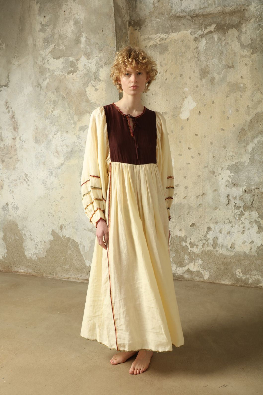 Model is wearing the Serafina Brown Linen Dress by Supernaturae x HONS, crafted in a blend of crisp linen and hand-spun silk with rich earthy botanical dye tones. Featuring a 1970s-inspired kaftan silhouette, hand-crochet edges and adjustable ties, this sustainable slow fashion piece shows natural tonal variations unique to each garment. Made in small-batch production in Mexico using ancestral techniques, part of the House of Woven Stories selection.
