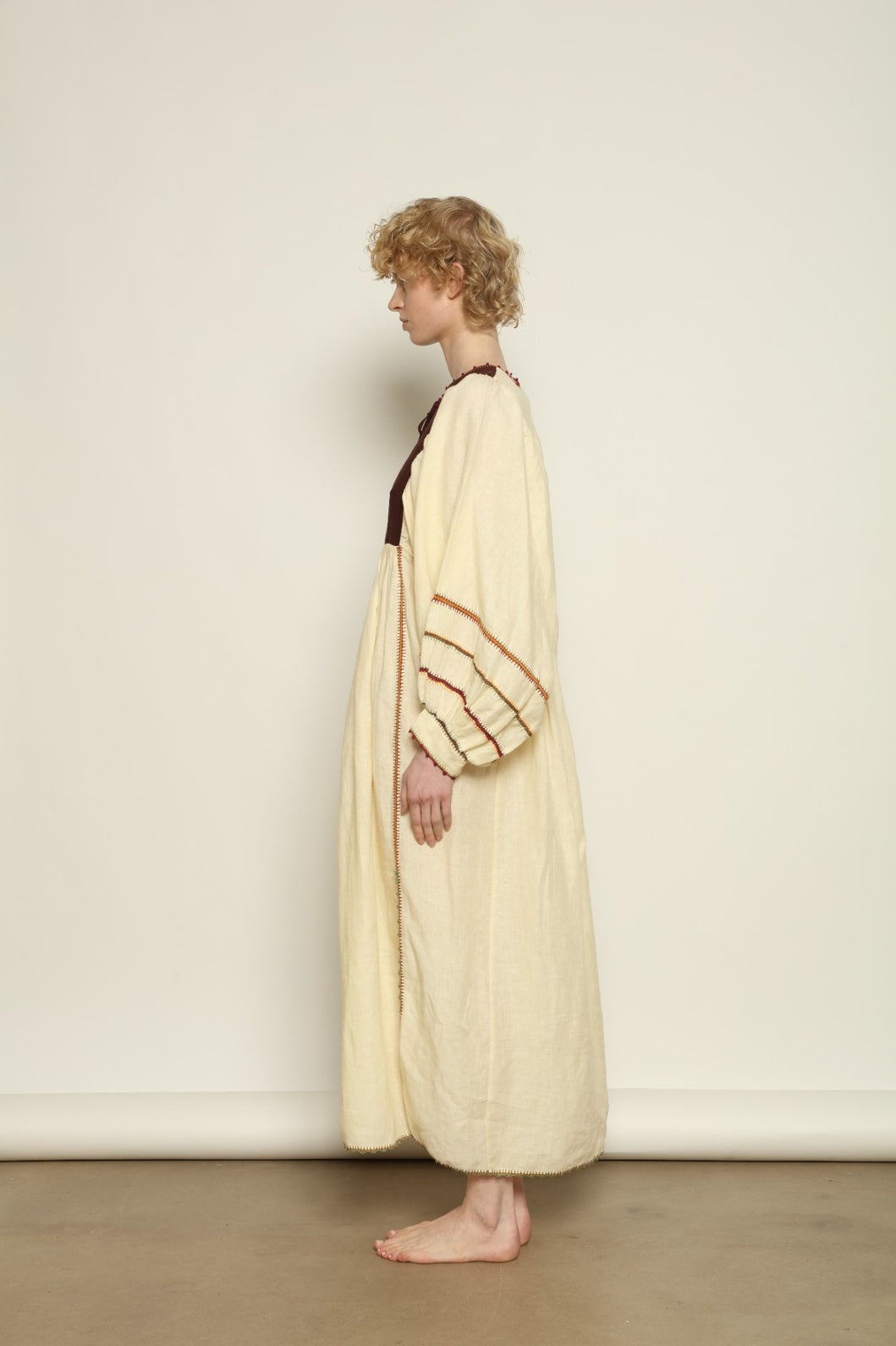 Model is wearing the Serafina Brown Linen Dress by Supernaturae x HONS, crafted in a blend of crisp linen and hand-spun silk with rich earthy botanical dye tones. Featuring a 1970s-inspired kaftan silhouette, hand-crochet edges and adjustable ties, this sustainable slow fashion piece shows natural tonal variations unique to each garment. Made in small-batch production in Mexico using ancestral techniques, part of the House of Woven Stories selection.