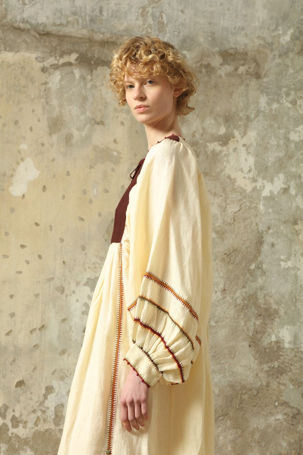 Model is wearing the Serafina Brown Linen Dress by Supernaturae x HONS, crafted in a blend of crisp linen and hand-spun silk with rich earthy botanical dye tones. Featuring a 1970s-inspired kaftan silhouette, hand-crochet edges and adjustable ties, this sustainable slow fashion piece shows natural tonal variations unique to each garment. Made in small-batch production in Mexico using ancestral techniques, part of the House of Woven Stories selection.