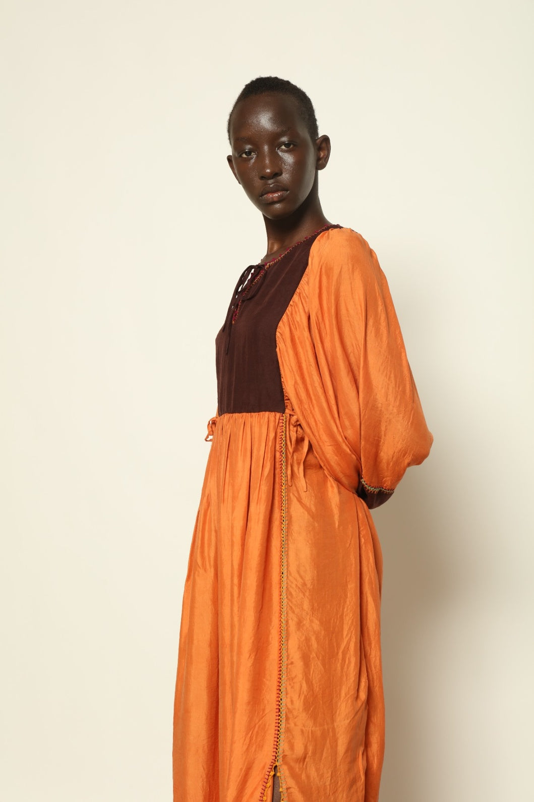 Model is wearing the Serafina Cognac Silk Dress by Supernaturae x HONS in warm cognac-toned handwoven silk, featuring a flowing kaftan-inspired silhouette and soft natural drape. Made with traditional artisan techniques and natural dyeing, this sustainable luxury dress has subtle tonal variations and adjustable side ties for a flexible fit, part of the House of Woven Stories selection.