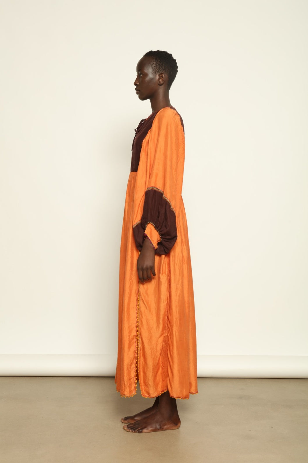 Model is wearing the Serafina Cognac Silk Dress by Supernaturae x HONS in warm cognac-toned handwoven silk, featuring a flowing kaftan-inspired silhouette and soft natural drape. Made with traditional artisan techniques and natural dyeing, this sustainable luxury dress has subtle tonal variations and adjustable side ties for a flexible fit, part of the House of Woven Stories selection.