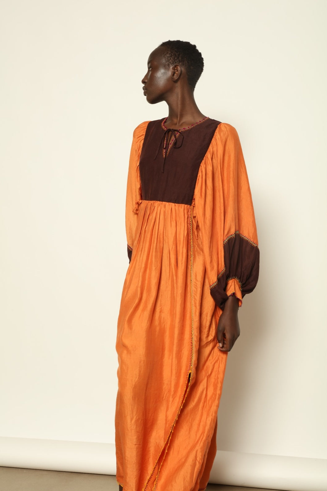 Model is wearing the Serafina Cognac Silk Dress by Supernaturae x HONS in warm cognac-toned handwoven silk, featuring a flowing kaftan-inspired silhouette and soft natural drape. Made with traditional artisan techniques and natural dyeing, this sustainable luxury dress has subtle tonal variations and adjustable side ties for a flexible fit, part of the House of Woven Stories selection.
