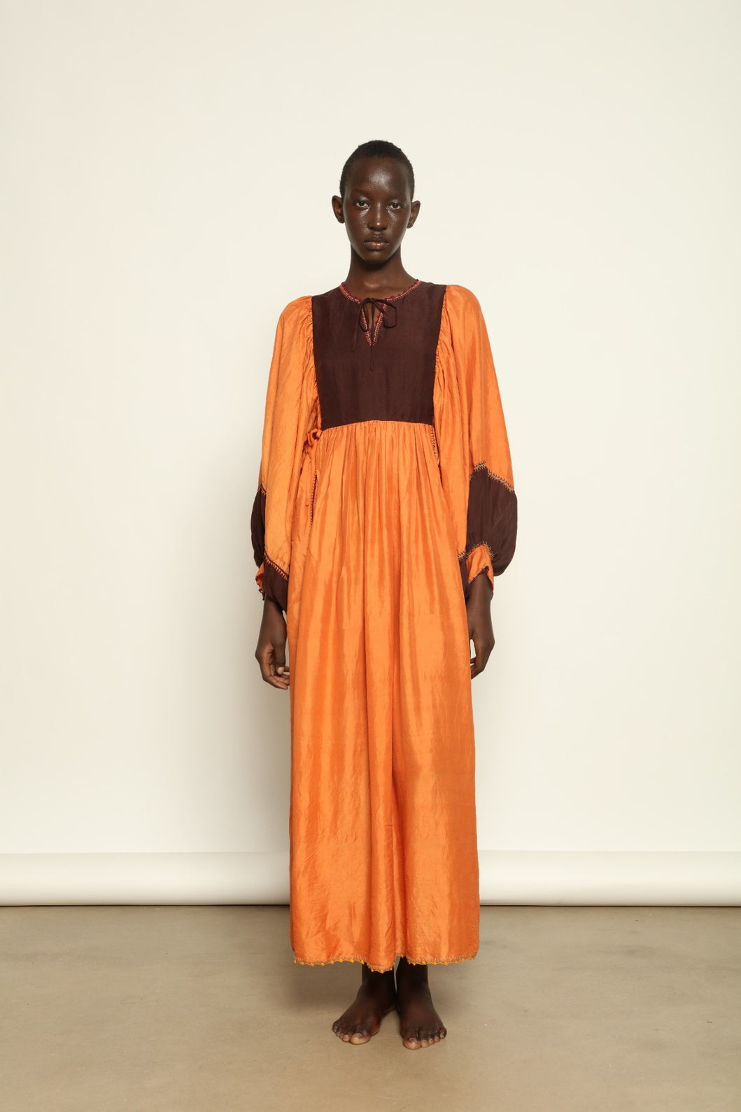 Model is wearing the Serafina Cognac Silk Dress by Supernaturae x HONS in warm cognac-toned handwoven silk, featuring a flowing kaftan-inspired silhouette and soft natural drape. Made with traditional artisan techniques and natural dyeing, this sustainable luxury dress has subtle tonal variations and adjustable side ties for a flexible fit, part of the House of Woven Stories selection.